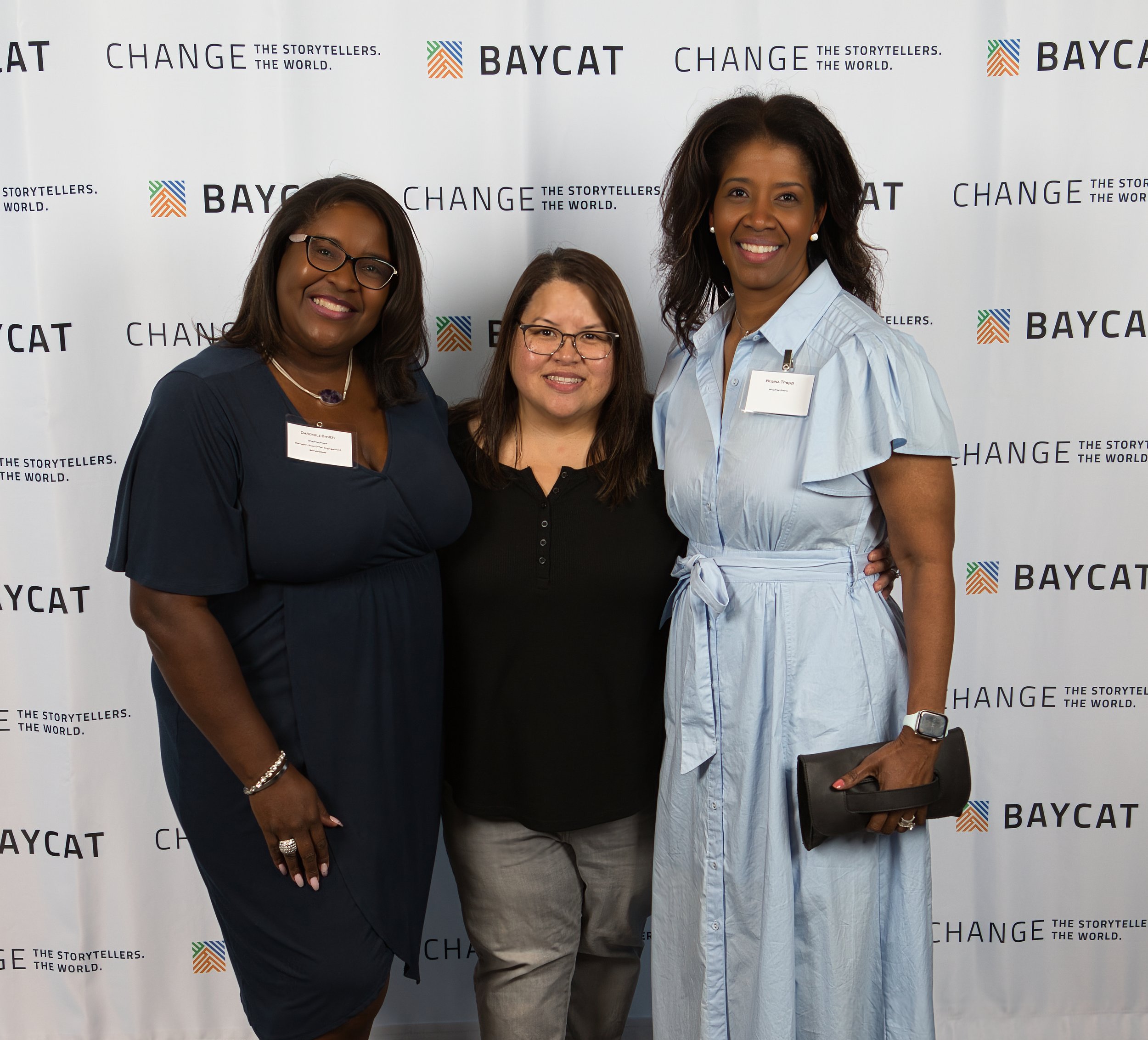 Three women standing together at an event with a backdrop that has the words 'BAYCAT,' 'CHANGE,' and the slogan 'The storytellers. The world.'