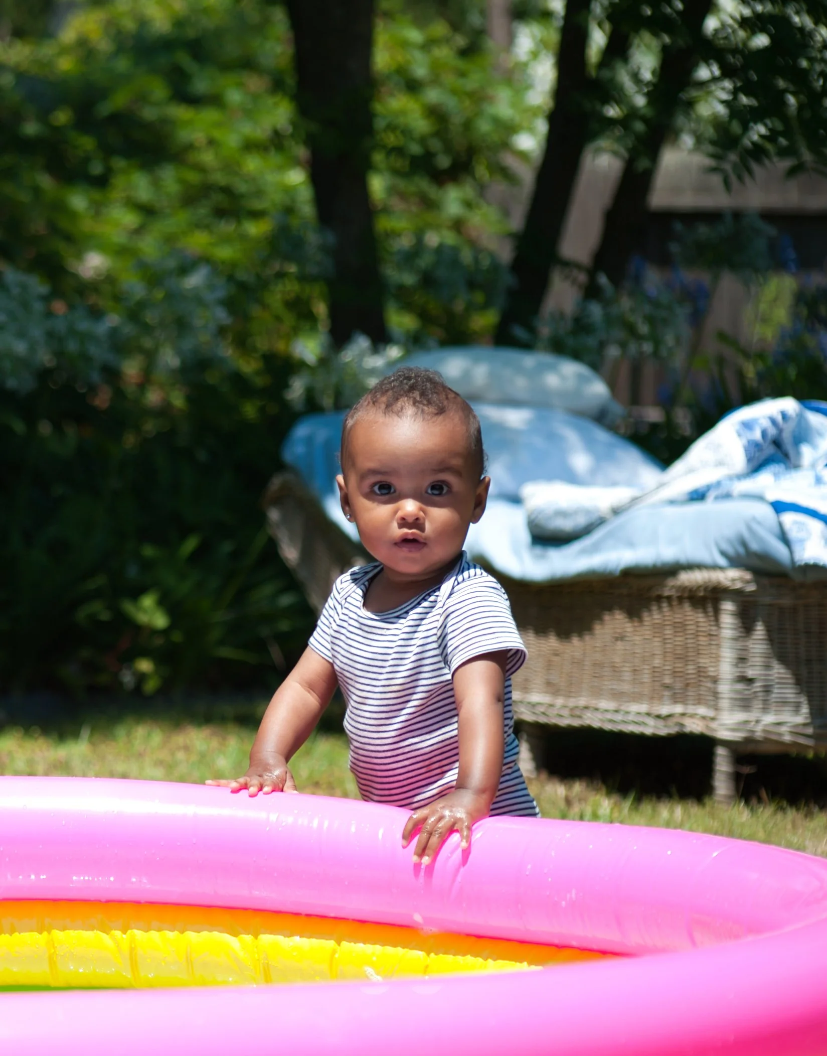 Baby standing in a pink inflatable pool outdoors on a sunny day, with trees and a wicker chair with a blue cloth in the background.