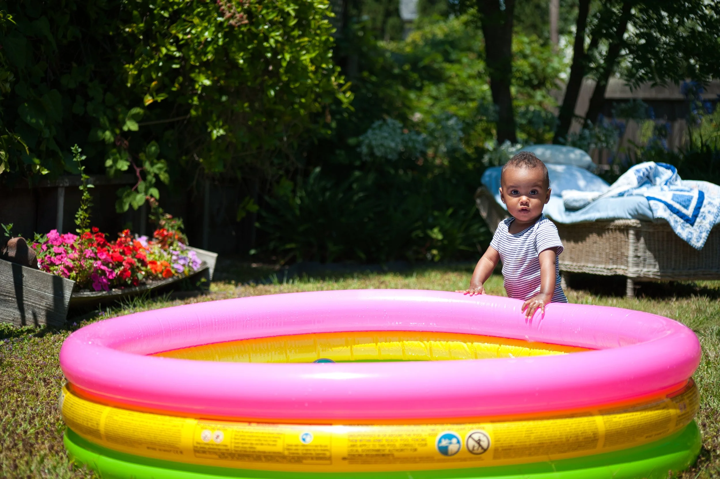 A young child standing in a colorful inflatable kiddie pool in a backyard with green grass, trees, and bushes. There is a wicker outdoor sofa with blankets in the background, and a flower bed with pink and purple flowers.