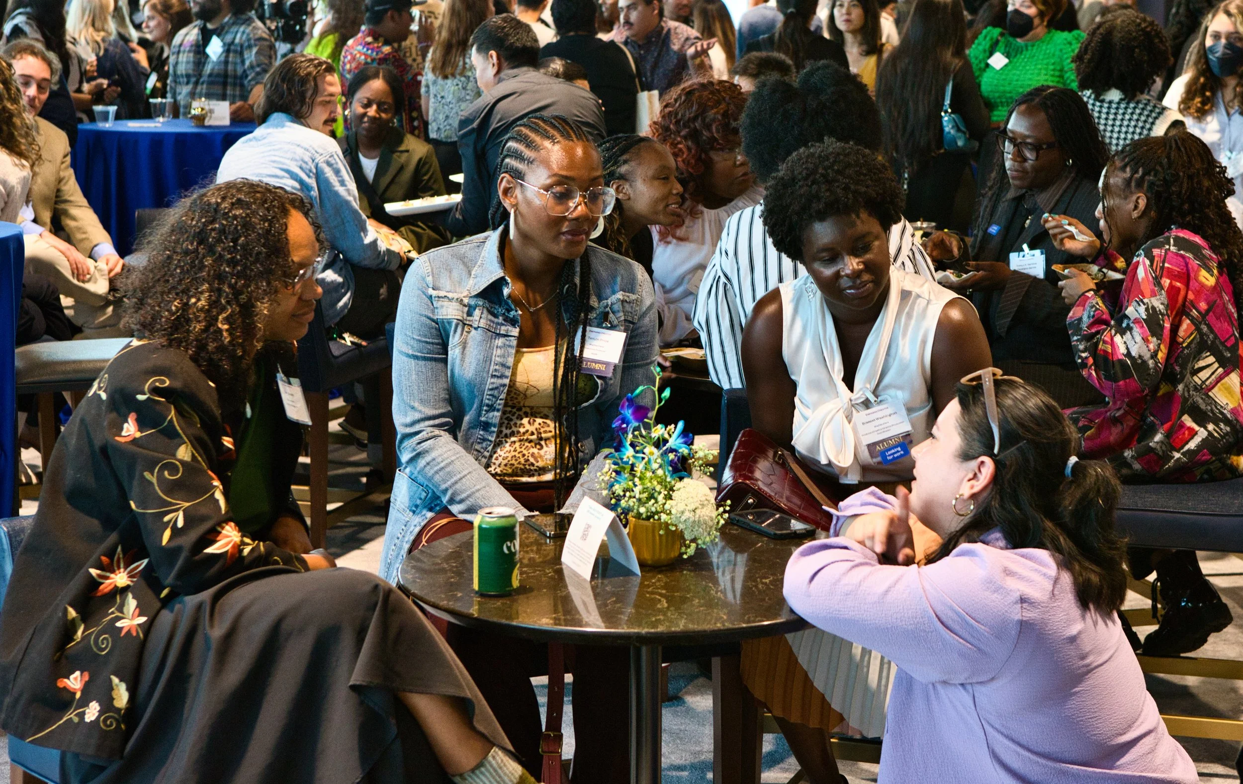 Group of women engaged in conversation at a networking or conference event, with many other attendees in the background.