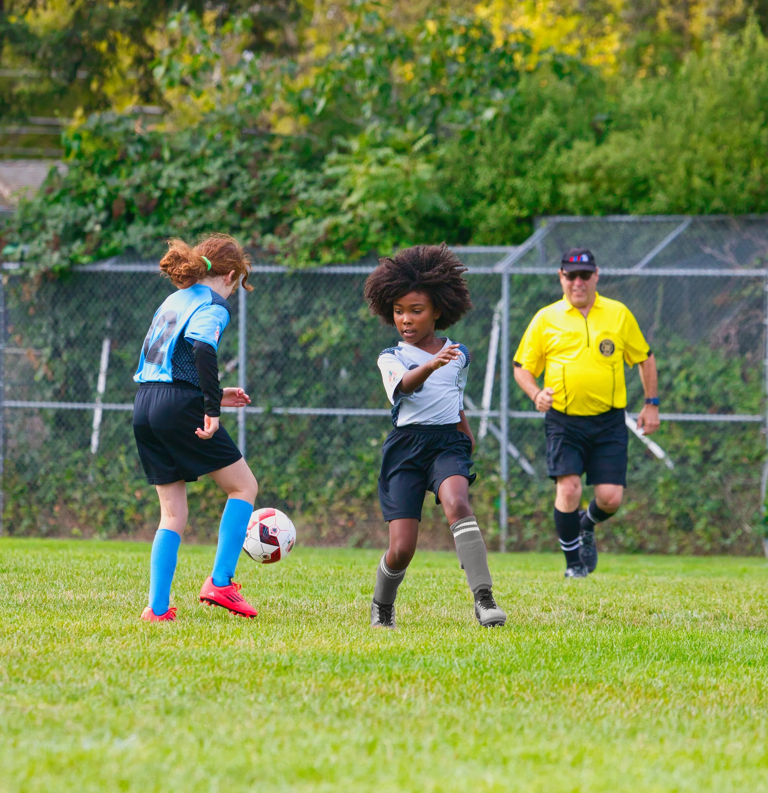 Two young girls playing soccer with a referee in the background on a grassy field surrounded by trees.