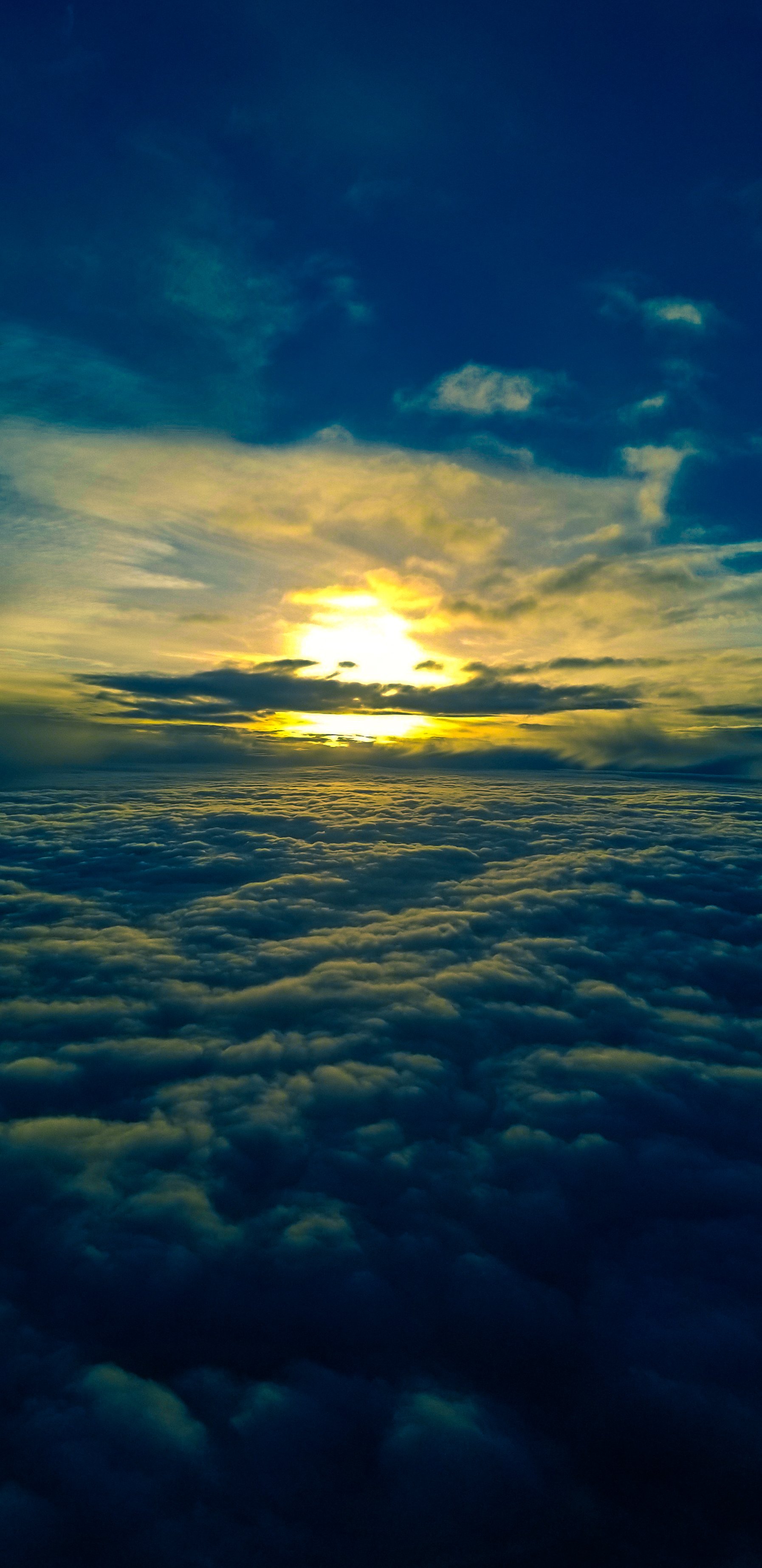 A view from above the clouds at sunrise or sunset, with the sky showing a gradient of blue to yellow, and the sun partially obscured by clouds.