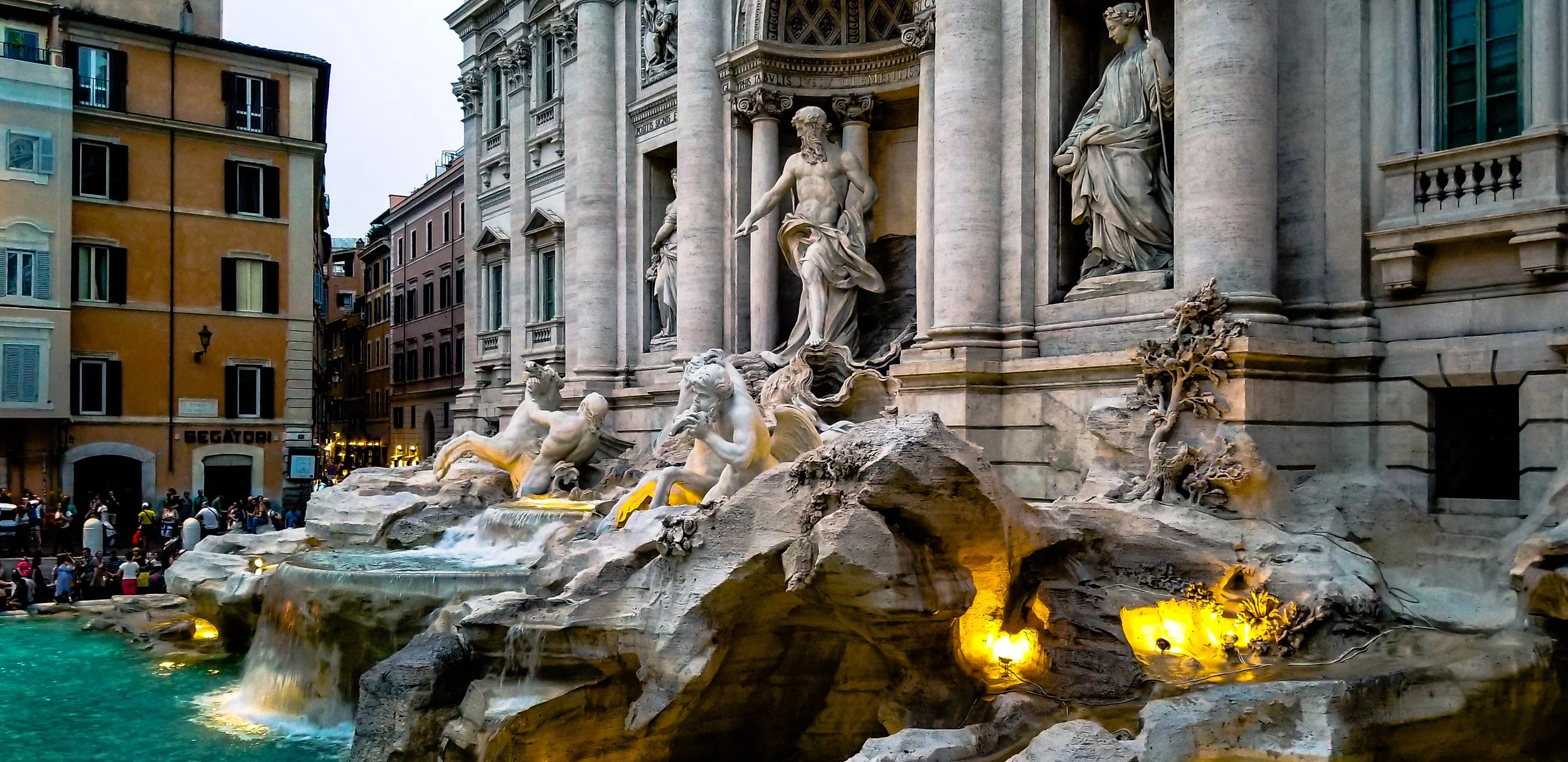 The Trevi Fountain in Rome, Italy, featuring intricate sculptures of gods, mythical creatures, and cascading water with surrounding buildings and tourists.