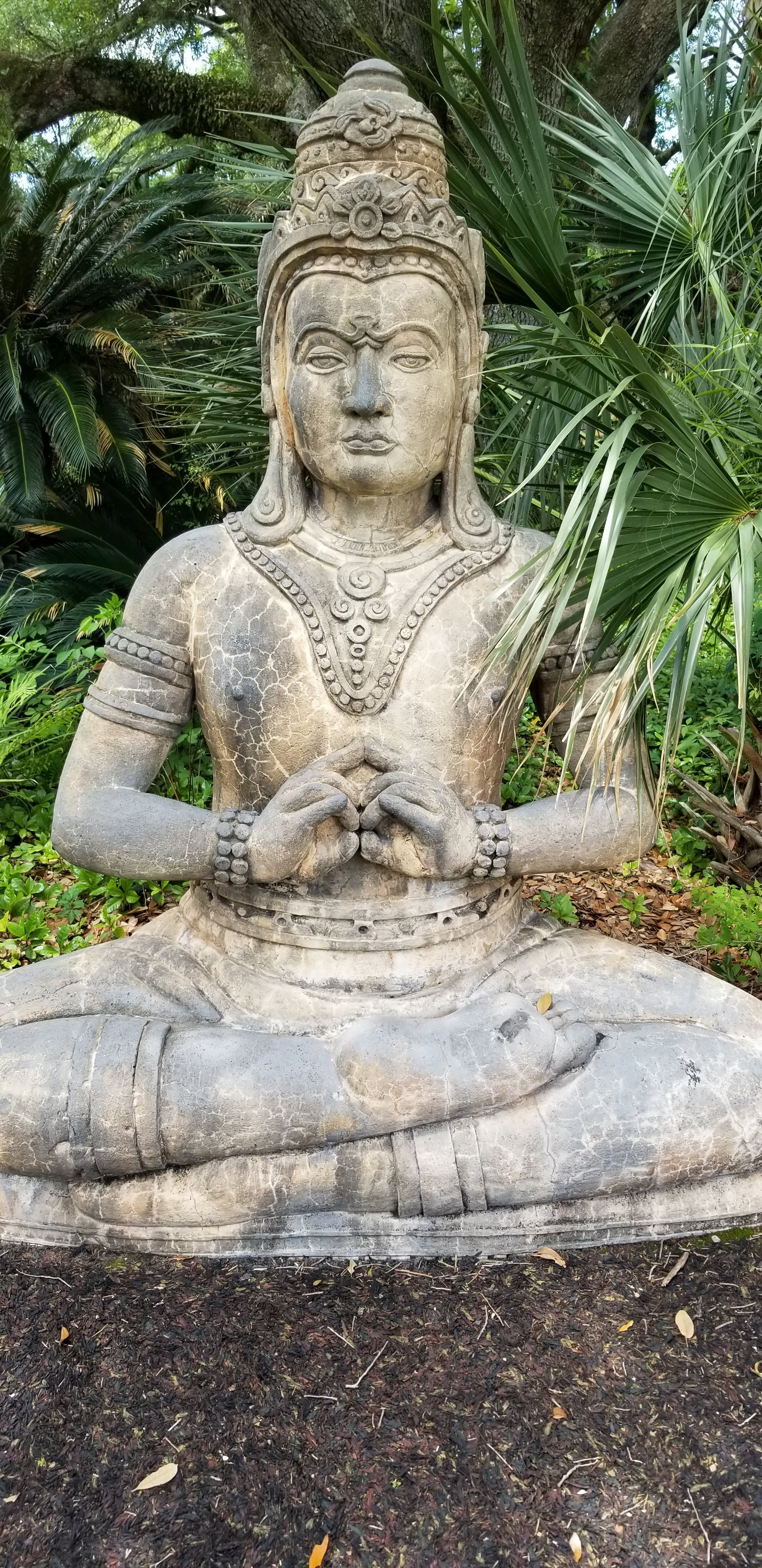 A stone statue of a seated person in a meditative pose, surrounded by lush green plants. The statue features detailed jewelry and a calm facial expression.