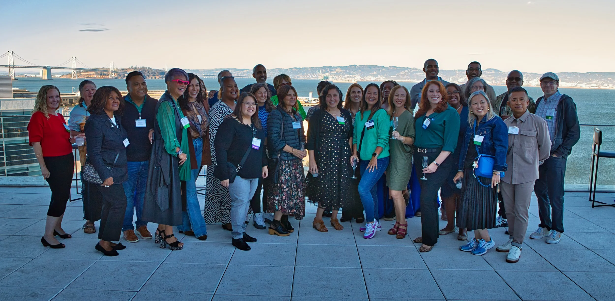 A diverse group of people gathered on a rooftop with a view of a bay and a bridge in the background, smiling for a photo during a social or professional event.