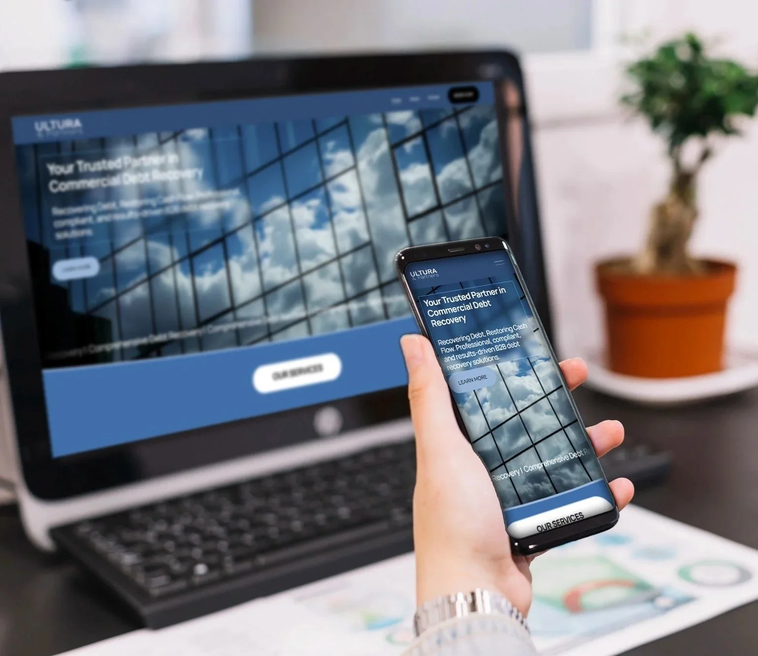 A person holds a smartphone displaying a webpage for a financial services company, with a laptop in the background showing the same webpage, and a potted plant in an orange pot on a white table.