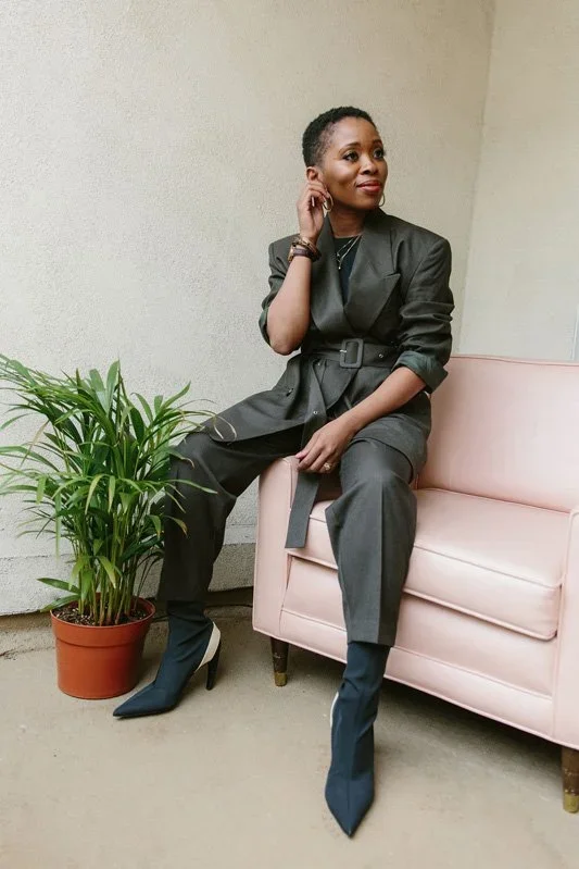 A woman in a dark gray suit sitting on a pink sofa next to a potted green plant, with short hair and a thoughtful expression.