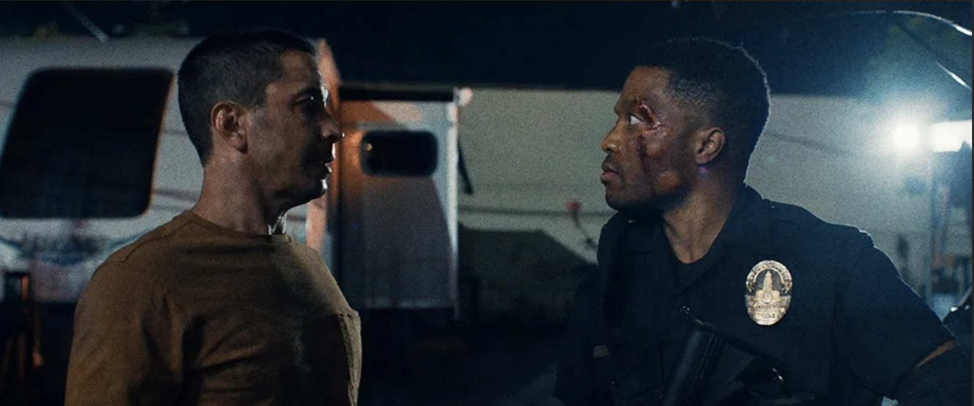 A tense confrontation between two men, one in a brown shirt and the other in a police uniform with a badge, inside a dimly lit space.