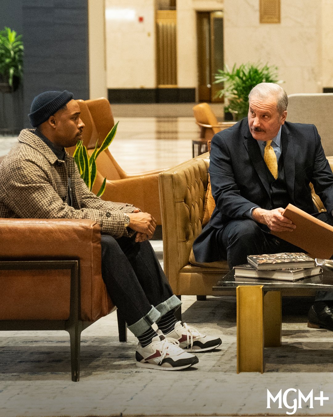 Two men sitting and talking in a hotel lobby, one dressed casually and the other in a suit, with a coffee table and magazines between them.