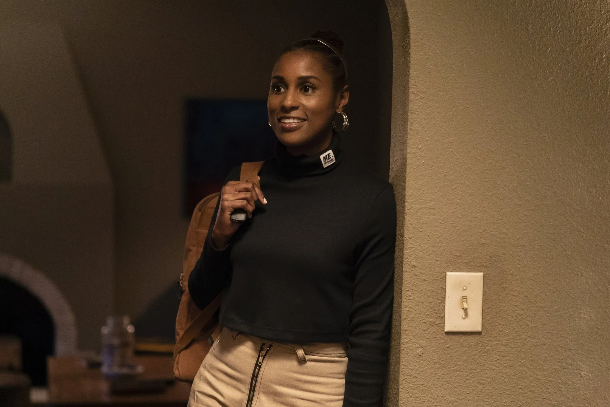 A young woman with dark hair tied back, wearing hoop earrings, a black turtleneck with a 'ME.' label, and beige pants, standing in an indoor hallway with a backpack over her shoulder, smiling and looking to her left.