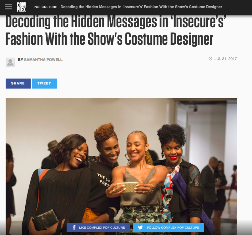 Four women are standing close together, smiling and looking at a smartphone, at an indoor event with a light background. They are dressed stylishly, and other people are visible in the background.