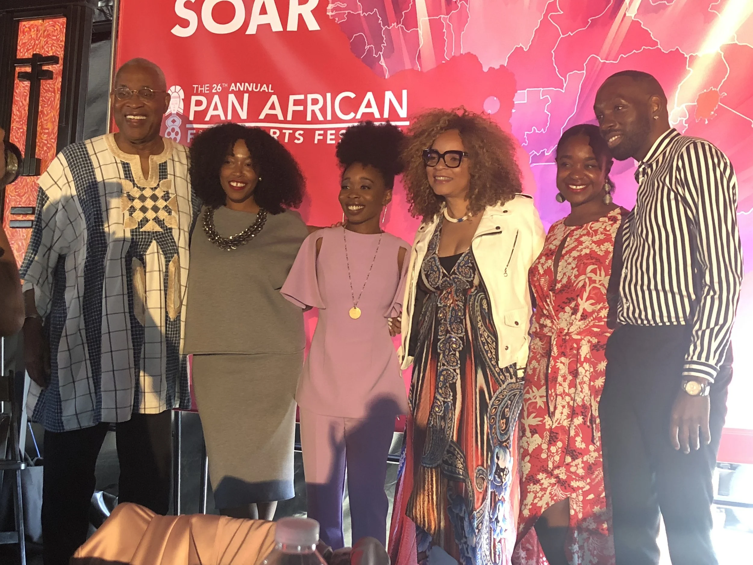 Group of six people standing on stage in front of a red backdrop at the 26th Annual Pan African Sports Festival, with a partially visible water bottle and bag in the foreground.