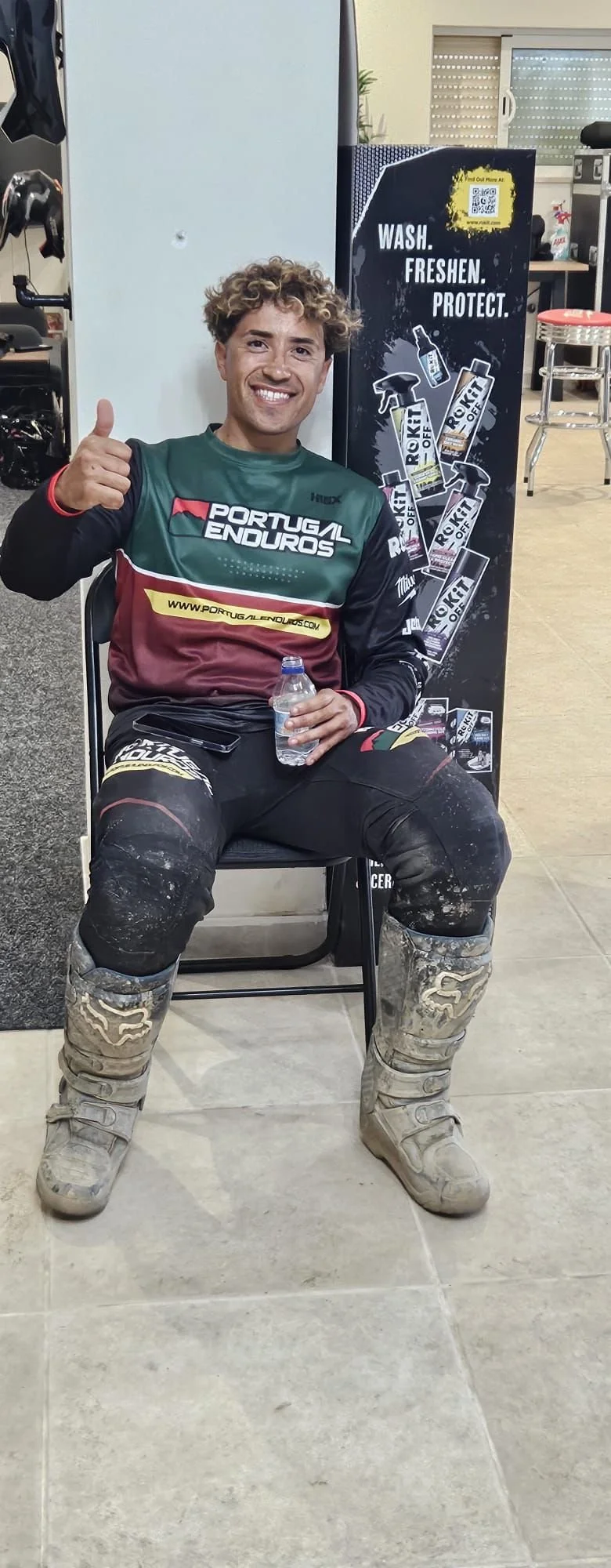 A smiling man wearing motocross gear sitting in a chair, giving a thumbs up, holding a water bottle, with a dirt-stained outfit and boots, sitting next to a vending machine that reads "Wash. Freshen. Protect." in a casual indoor setting.