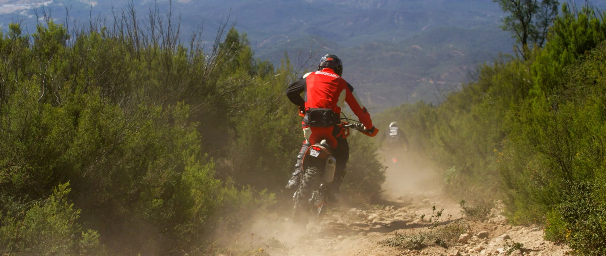 Two motorcyclists riding dirt bikes on a dusty trail through a mountainous landscape with green bushes and trees.