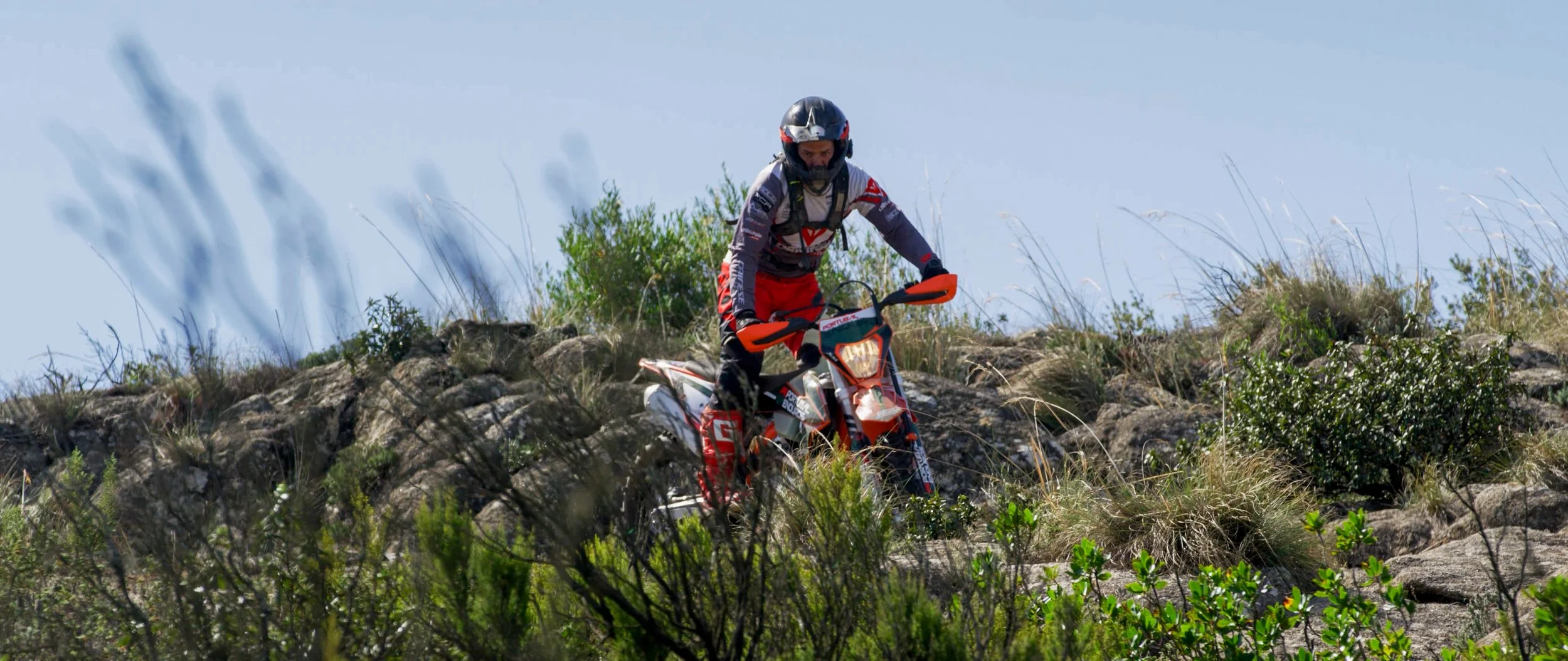 Motorcycle rider navigating rocky trail surrounded by shrubbery, wearing a helmet and protective gear.