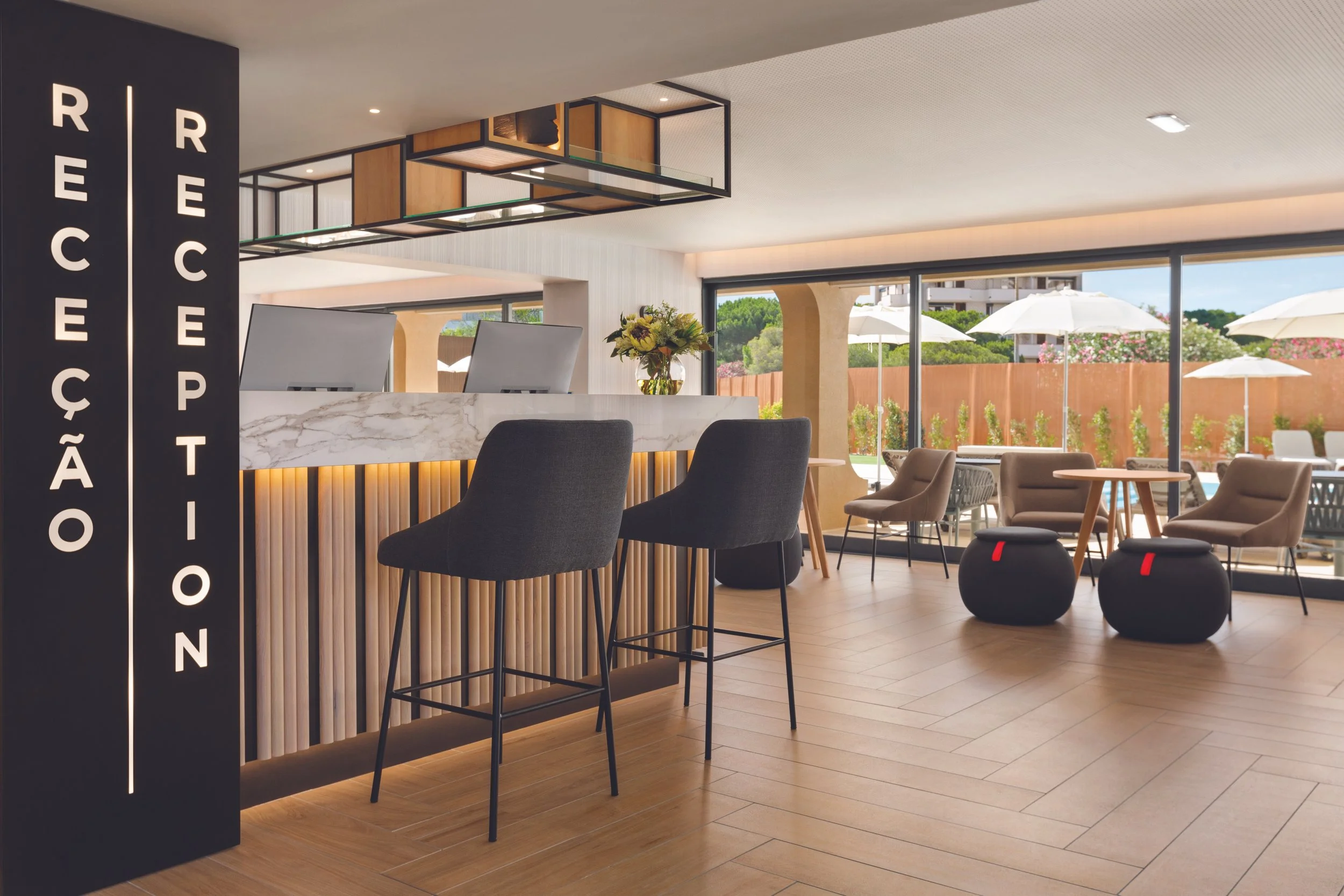 Modern hotel reception area with a marble-topped desk, high chairs, seating area with armchairs and ottomans, large windows, and outdoor umbrellas