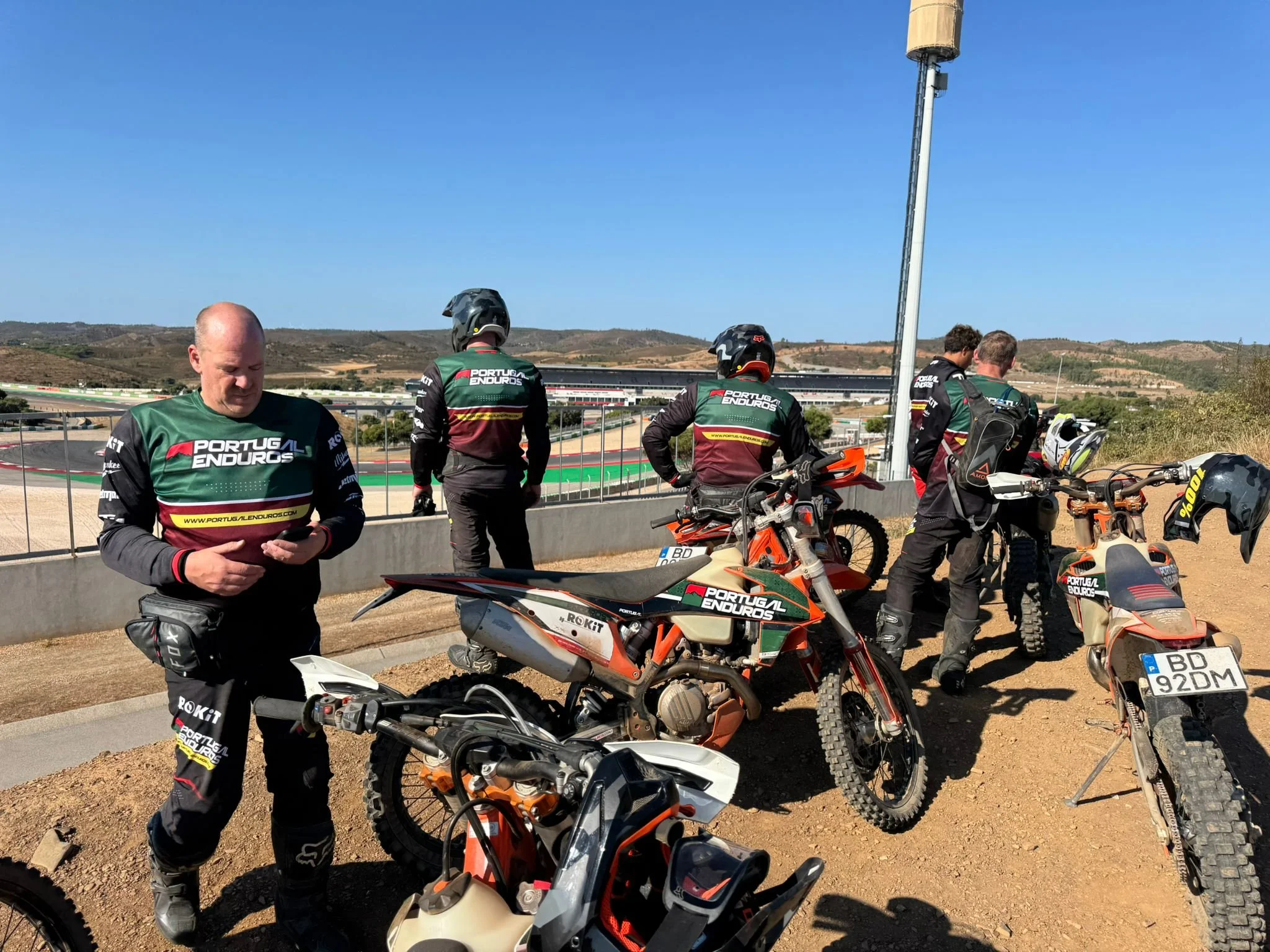 A group of five motorcyclists in Portugal Enduros gear are taking a break on a dirt track, with racing bikes parked nearby. They are outdoors on a sunny day, with a race track, hills, and stakes in the background.