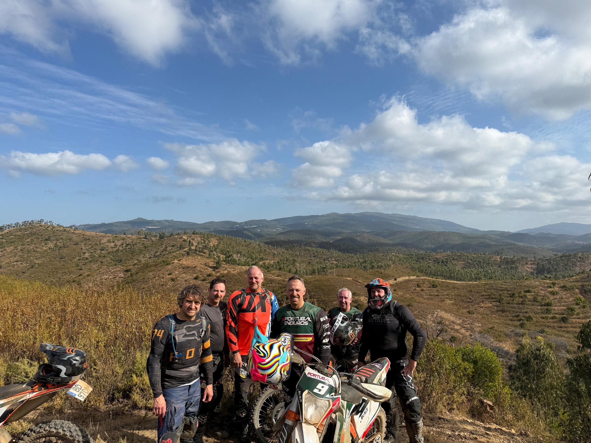 Group of six men standing outdoors on a dirt trail, with mountains and a partly cloudy sky in the background, some wearing motocross gear and holding bikes.