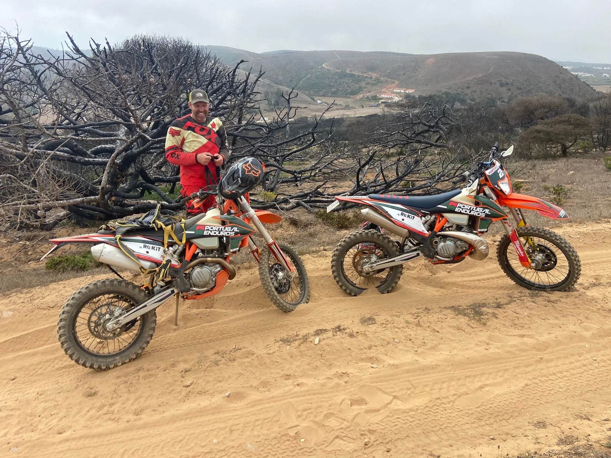 A man dressed in red and beige motocross gear standing next to two orange and white off-road dirt bikes on a sandy trail. There are leafless bushes and hills in the background under a cloudy sky.