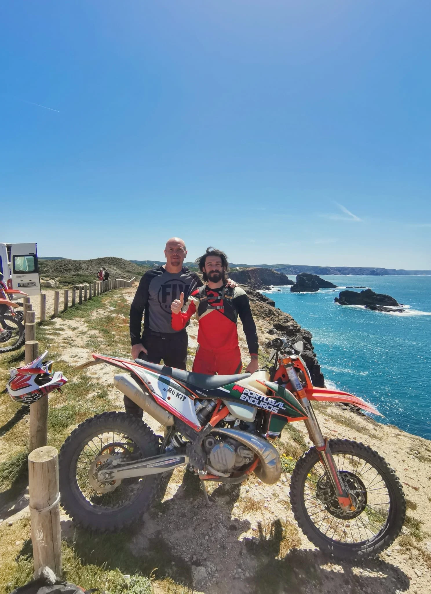 Two men standing next to a dirt bike on a coastal cliff with the ocean in the background, a dirt road, and a white vehicle.