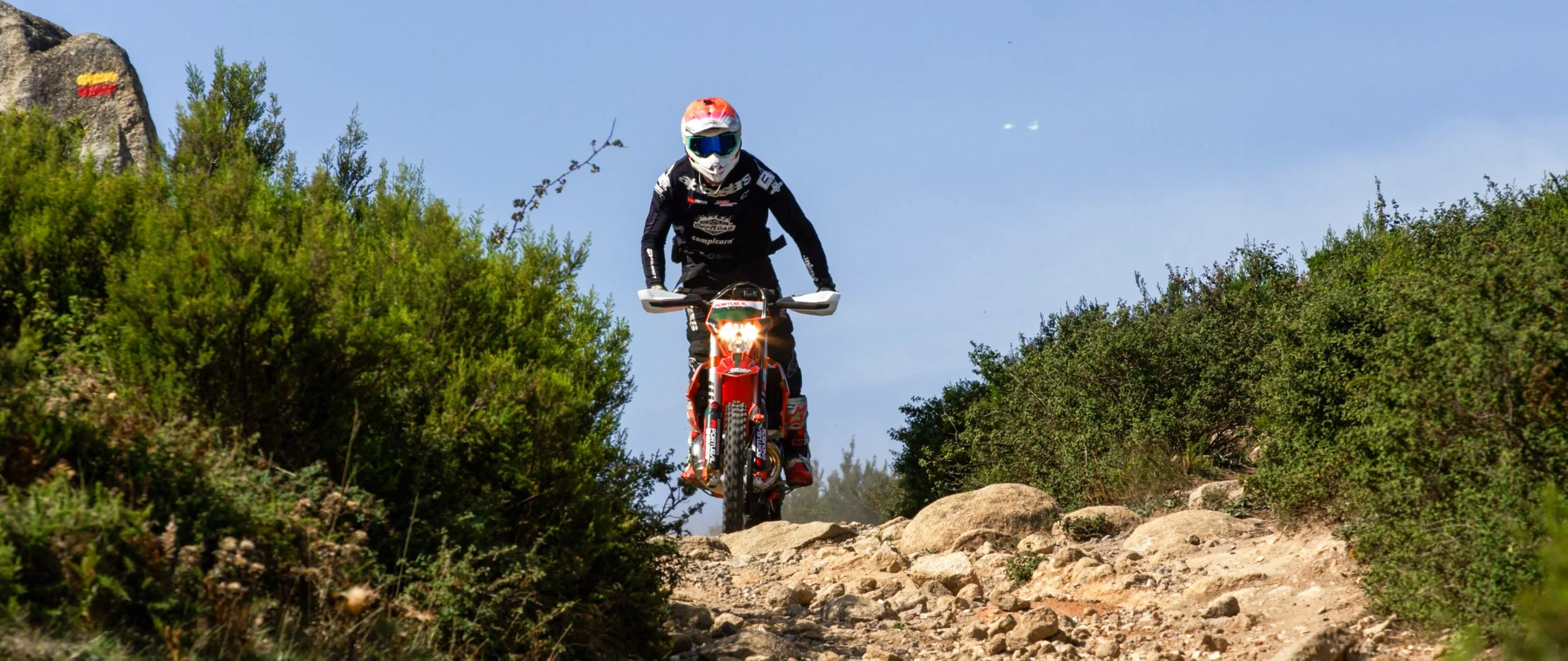 A motocross rider in black gear and a white helmet riding a dirt bike on a rocky trail surrounded by green bushes and trees under a clear blue sky.