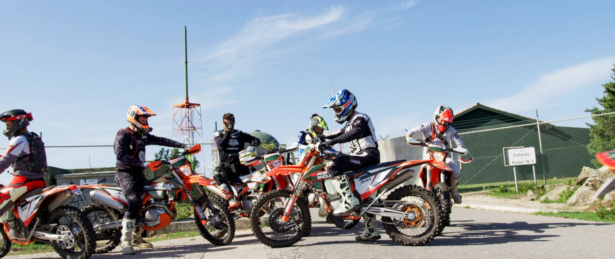 Group of motocross riders in gear with orange and white dirt bikes on a paved area outdoors under a blue sky.