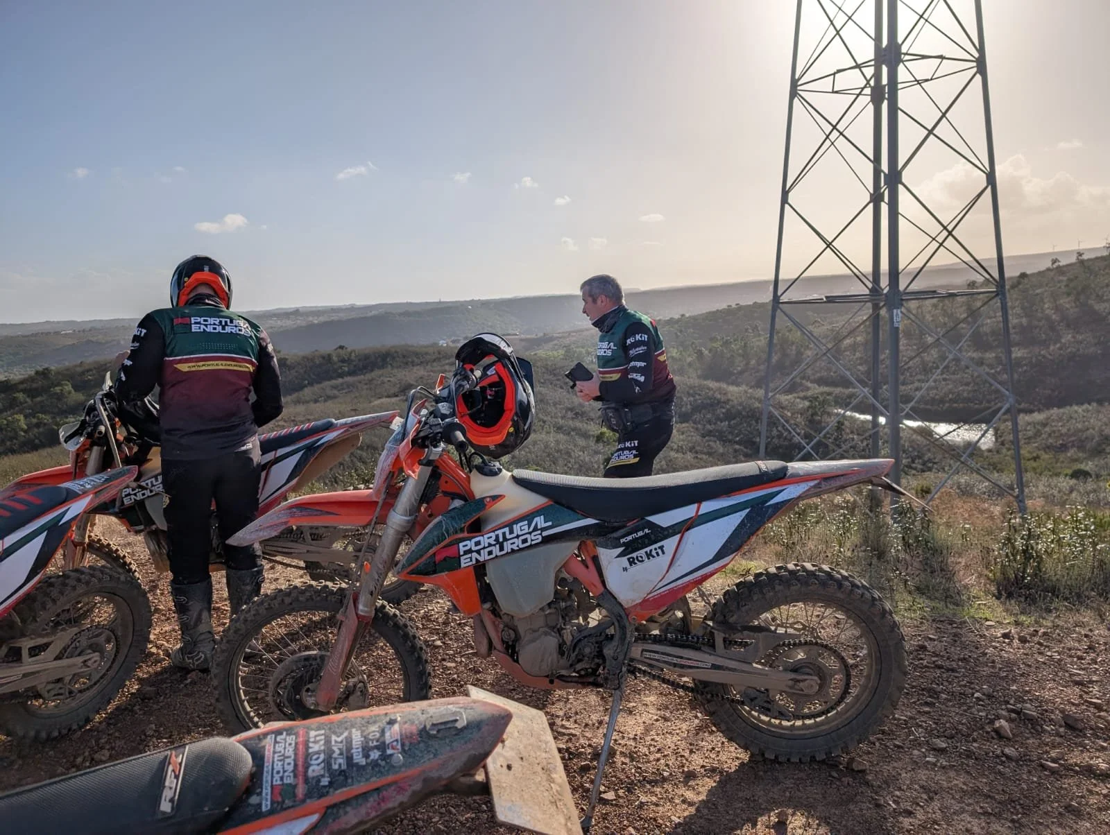 Two off-road motorcycles with Portugal Enduro racing decals parked on a dirt trail. Two men in racing gear are near the bikes, one facing away and the other looking at a device. The landscape has rolling hills under a partly cloudy sky, with a tall m