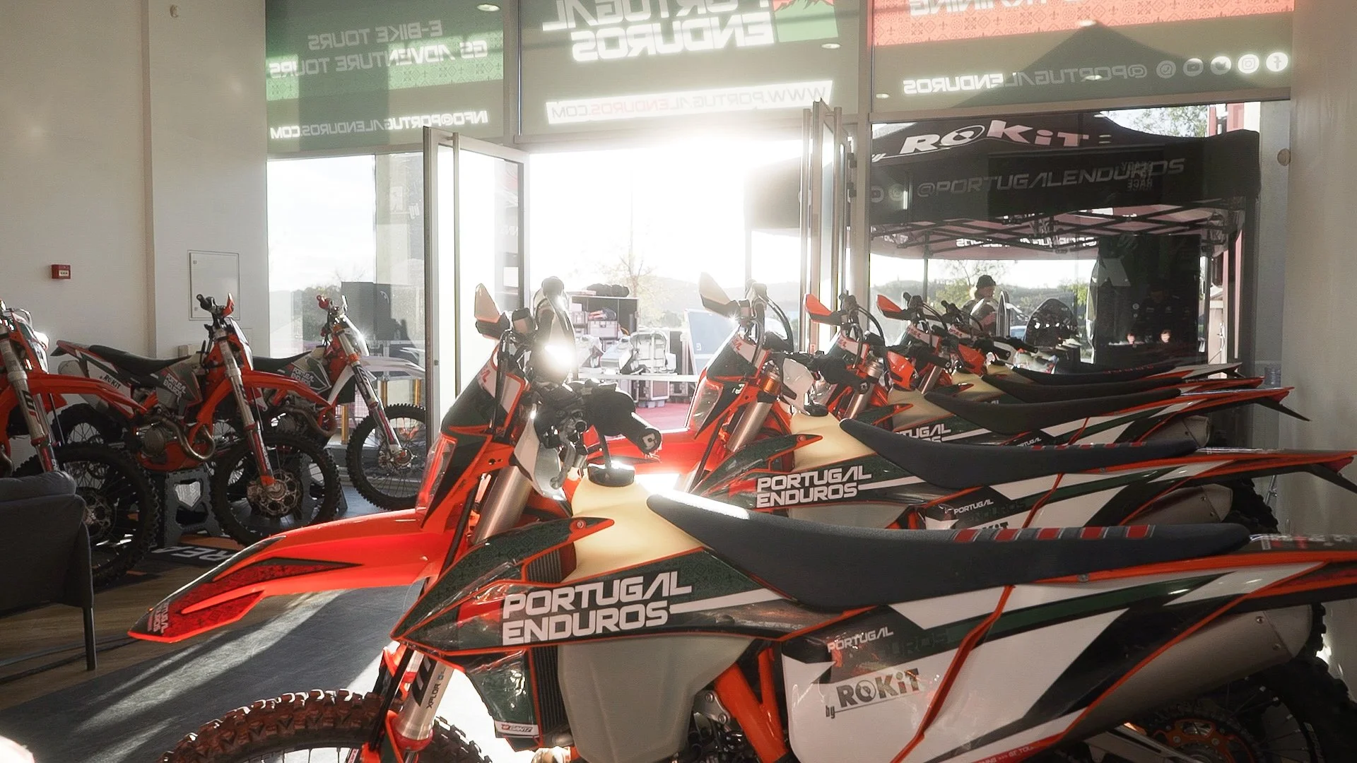Several Portugal Enduros motorcycles lined up inside a showroom, with more off-road bikes visible in the background. Bright sunlight streams in through the open glass entrance.