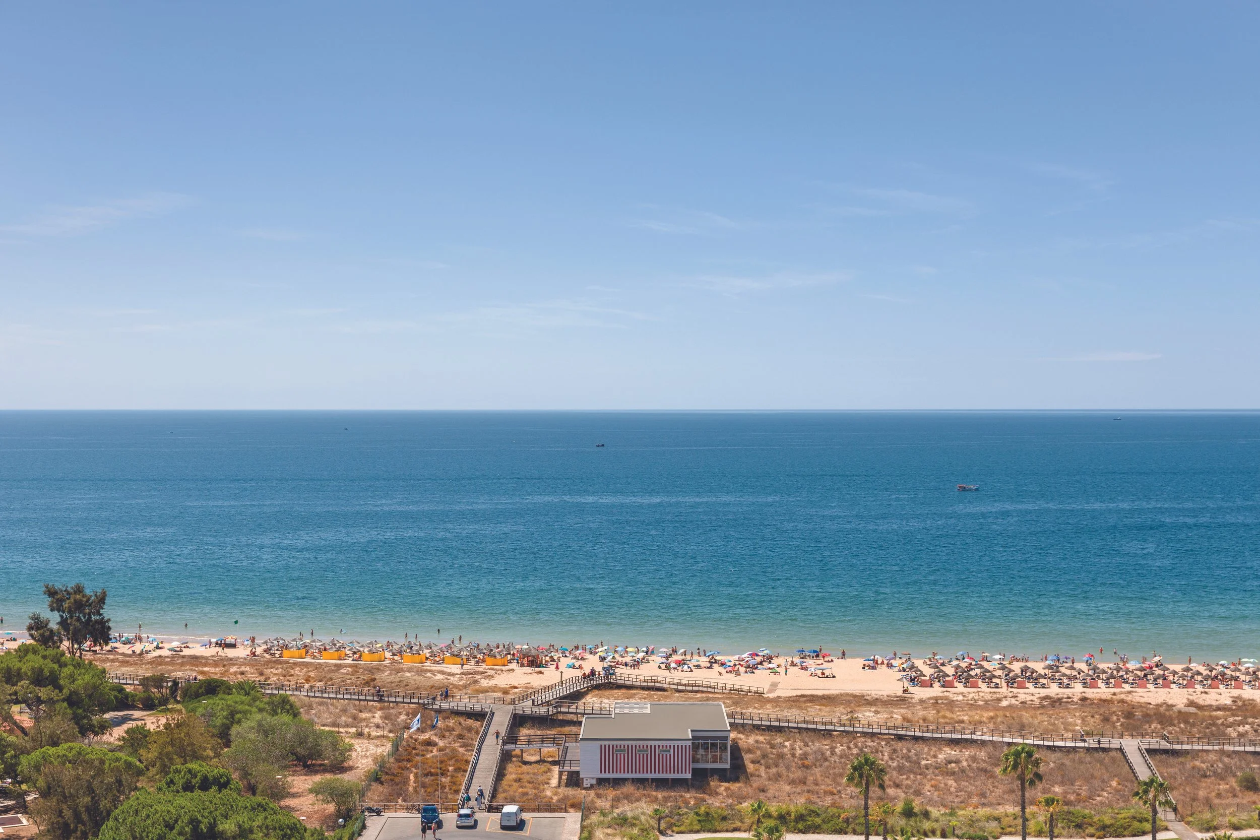 A beautiful beach scene with clear blue water and a sandy shoreline filled with colorful umbrellas and people. There is a small beach house, palm trees, and lush greenery in the foreground, with a clear sky above.