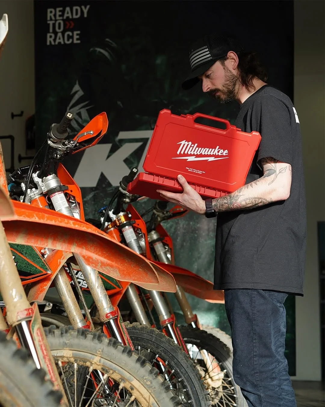 A man with a tattooed arm holding a red Milwaukee toolbox, standing next to off-road motorcycles at a dirt bike dealership or workshop.