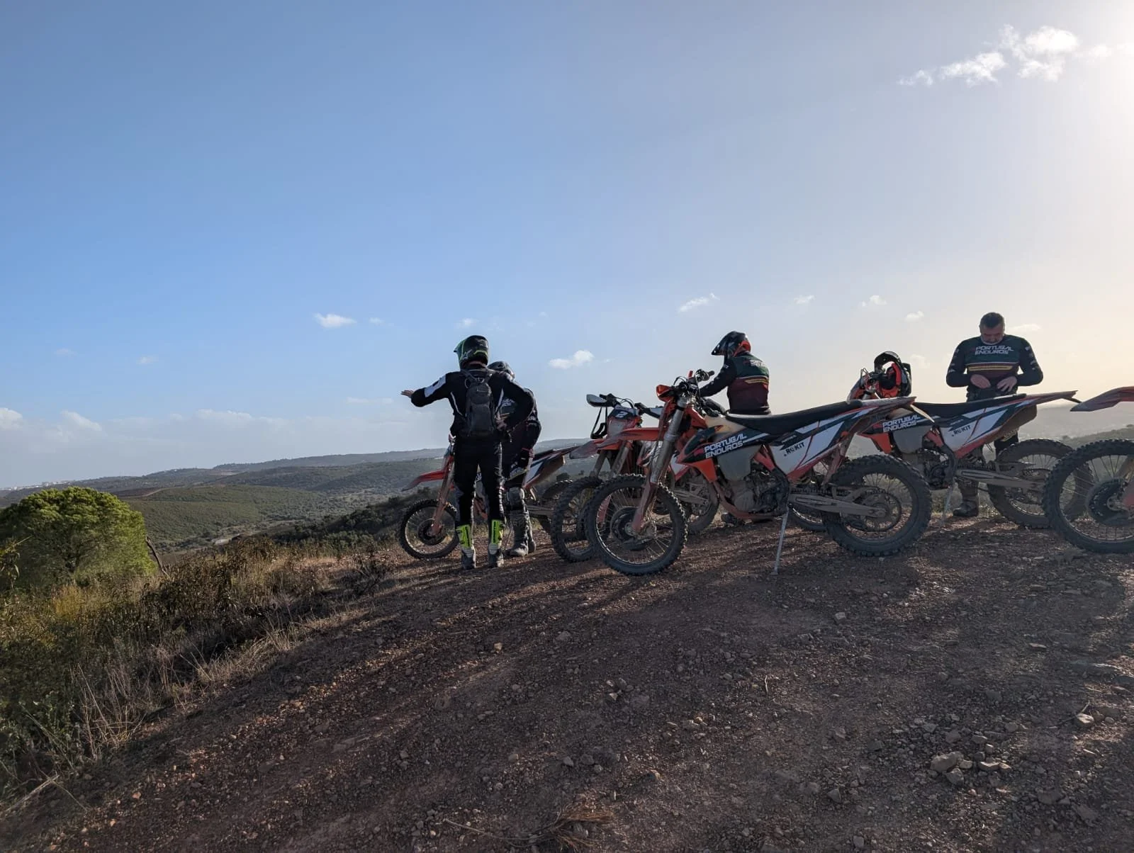 Group of motocross riders with off-road motorcycles on a dirt hill, overlooking a hilly landscape under a blue sky with some clouds.