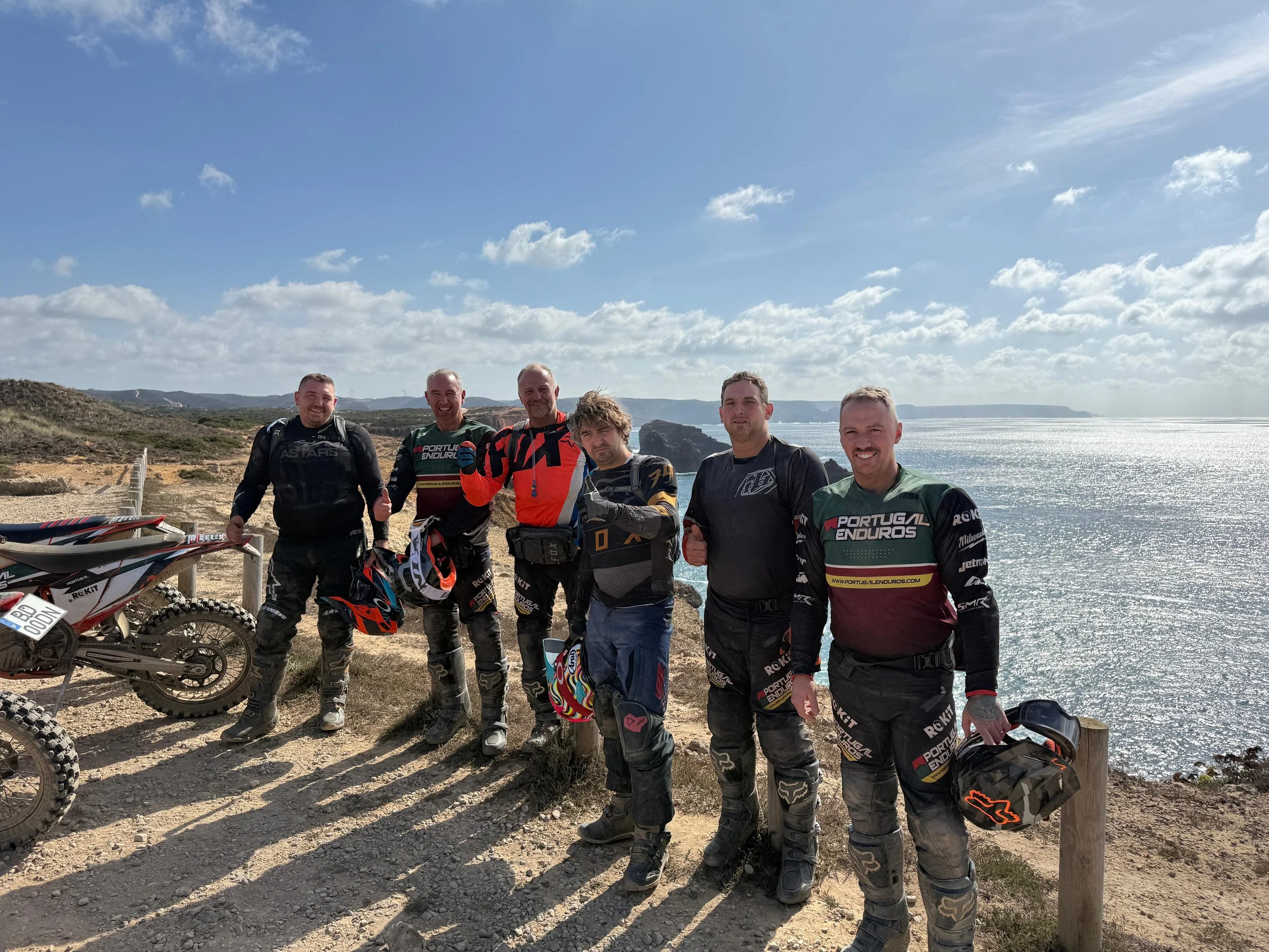 Six men in motocross gear standing on a dirt path near the ocean, with a motorcycle to the left, holding helmets, and an ocean and cloudy sky in the background.