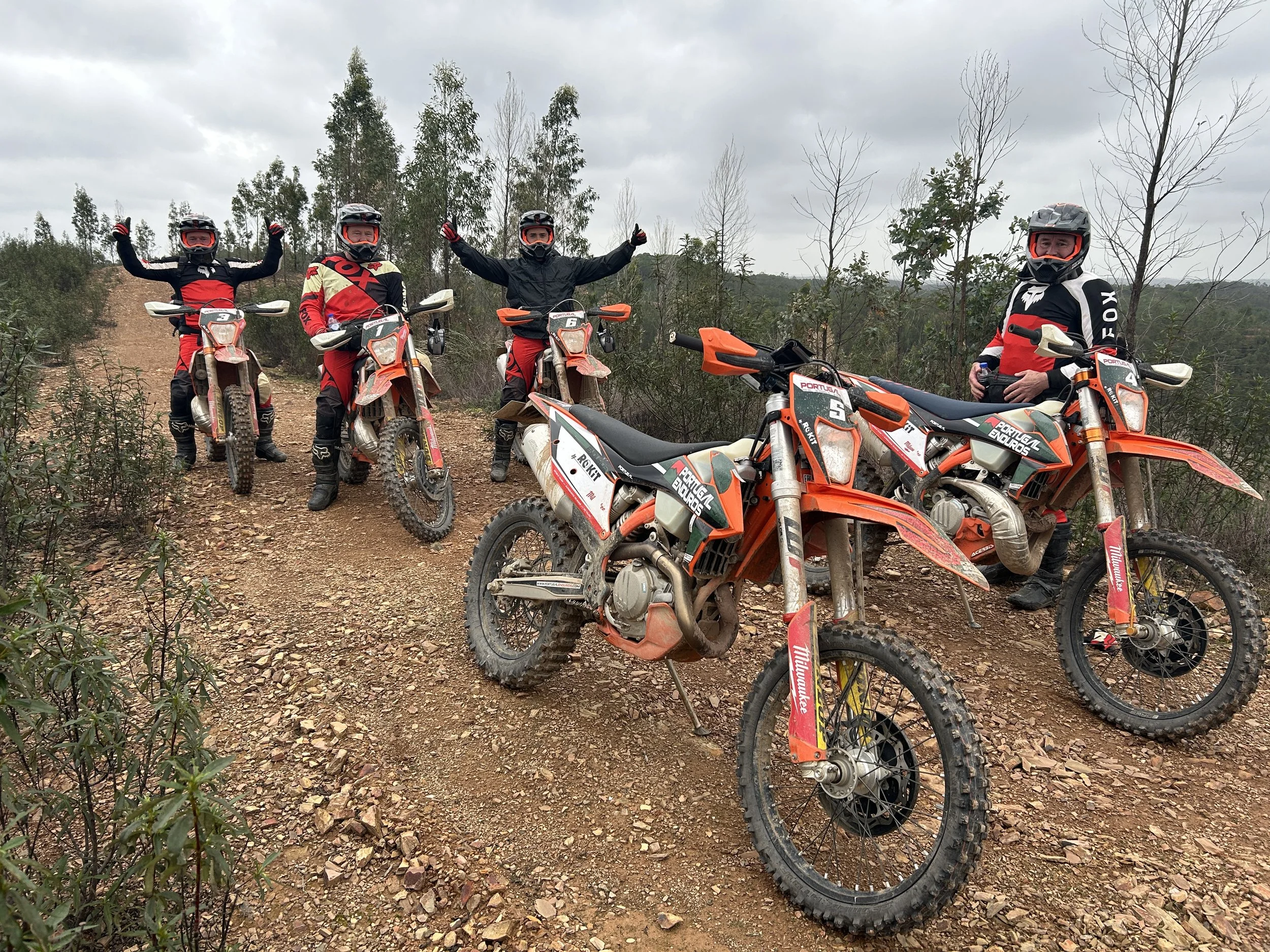 Four motocross riders in gear standing next to their dirt bikes on a dirt trail in a wooded area with cloudy sky.