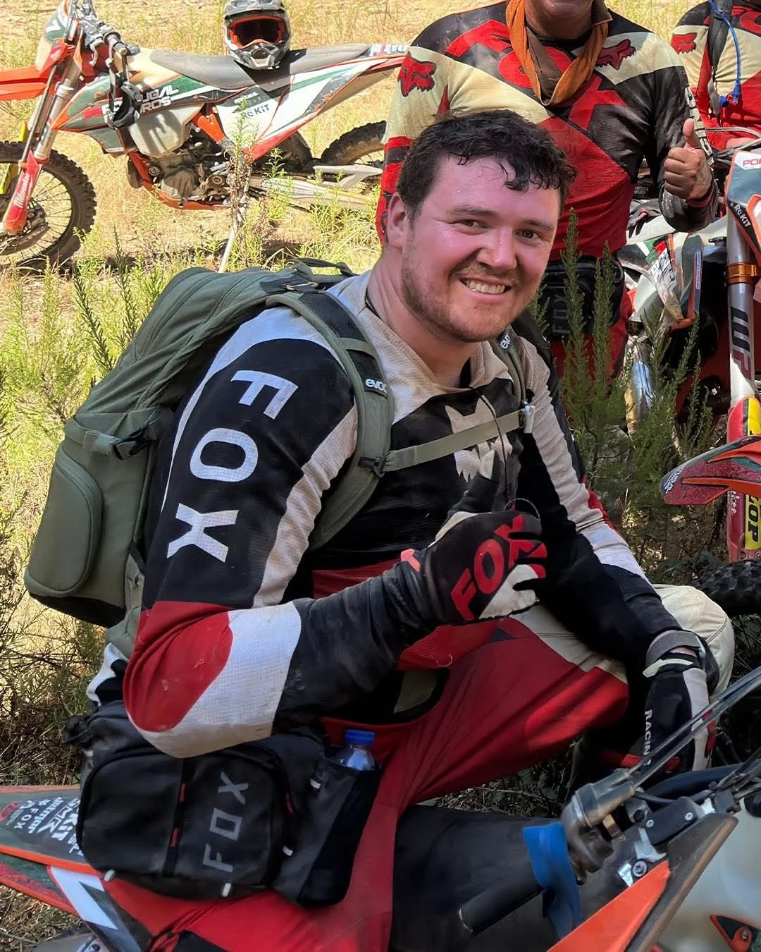 A man with a backpack and motocross gear smiling and sitting on a dirt bike, with other motocross bikes and riders in the background.