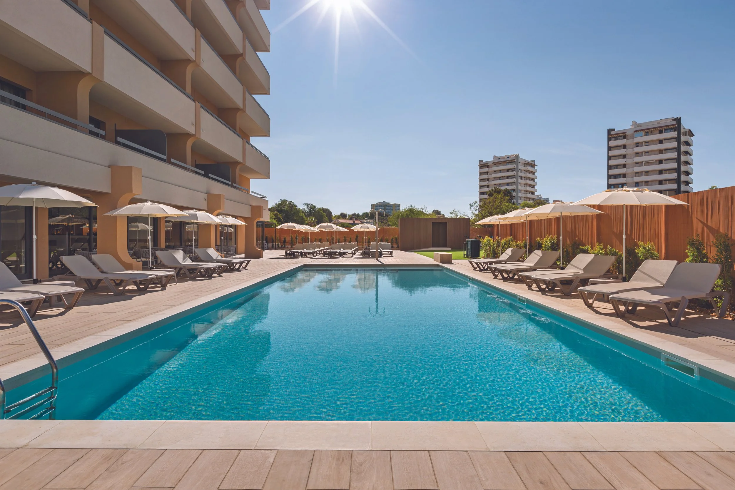 Outdoor swimming pool area with lounge chairs and umbrellas, surrounded by a modern residential building and high-rise apartments under a clear blue sky.
