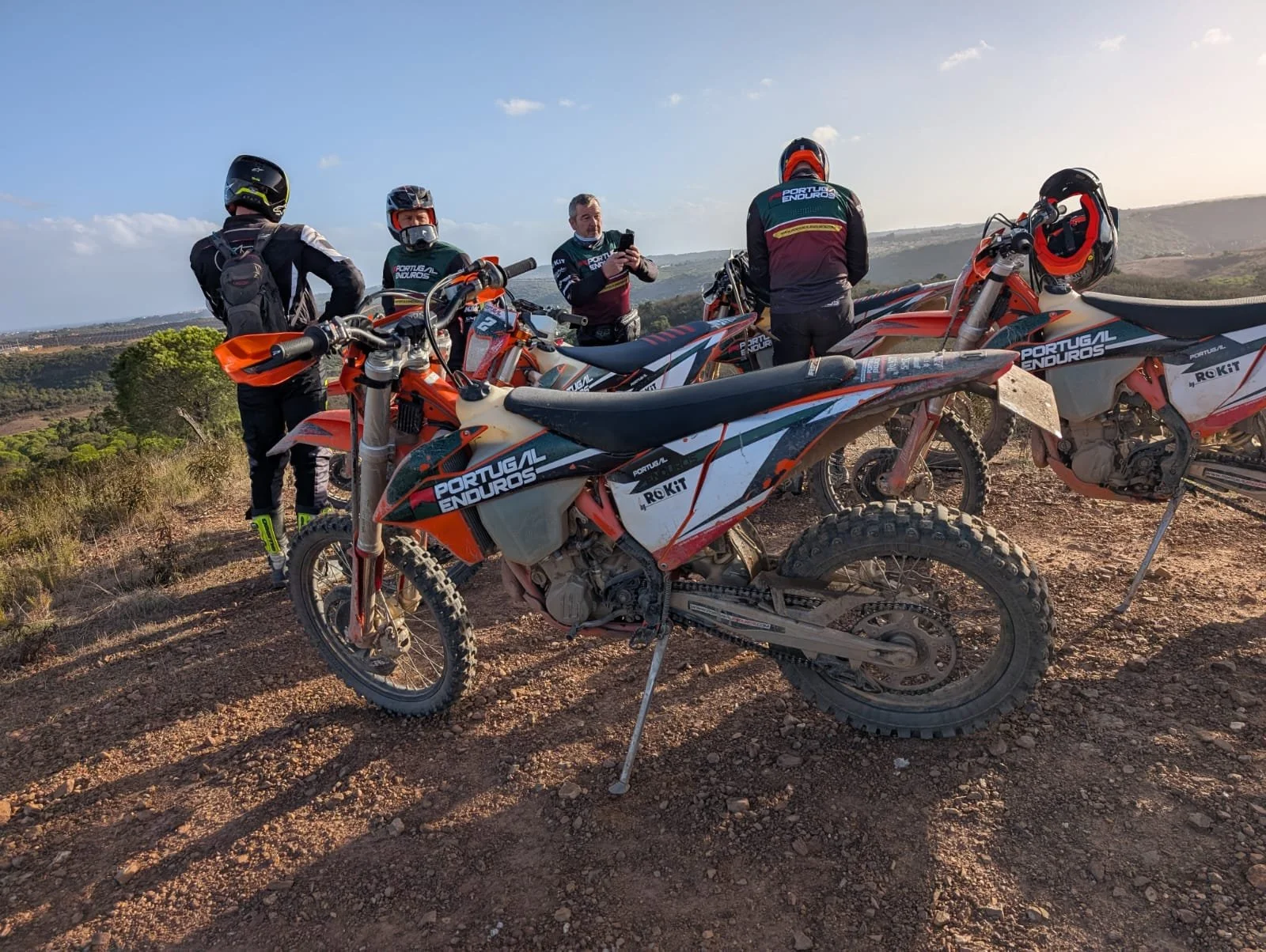 Group of four enduro motorcyclists taking a break on a dirt trail, with their bikes parked on a rocky hilltop against a landscape of rolling hills and blue sky.