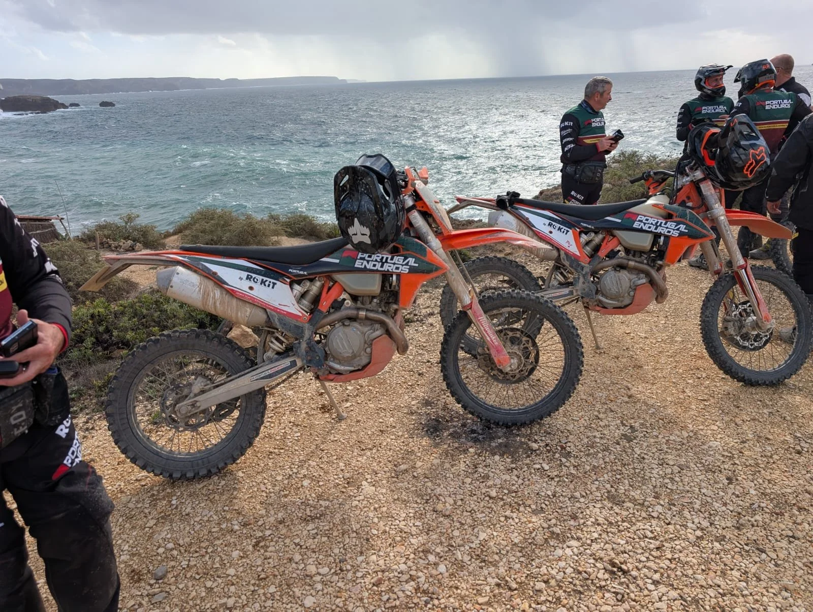 Two orange and black off-road motorcycles with dirt on them parked on a gravel trail near the ocean, with several people in motorcycle gear standing nearby, some looking at their phones, under partly cloudy skies.