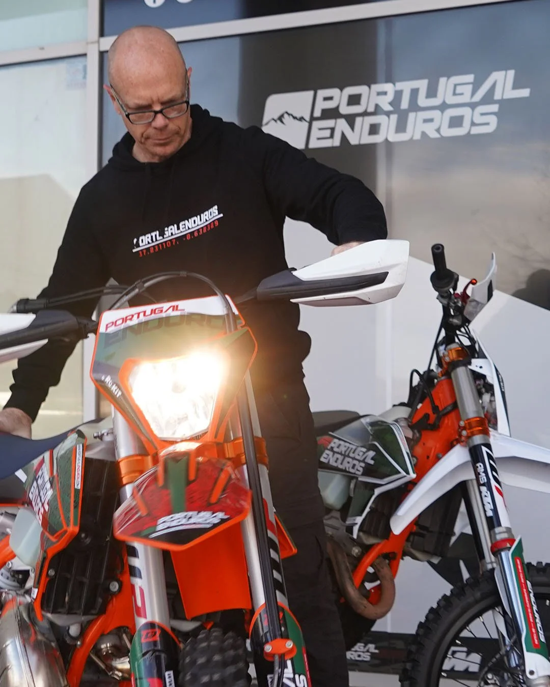 A man with glasses in black clothing is standing next to two orange and white off-road motorcycles, inspecting or working on one of them at a Portugal Enduros event.