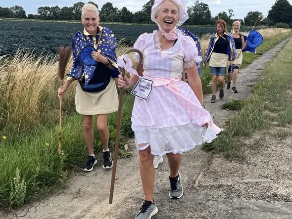 Group of four elderly women walking along a country path, wearing blue jackets with white star patterns, except one in a pink and white dress. They are smiling and appear to be enjoying a walk.