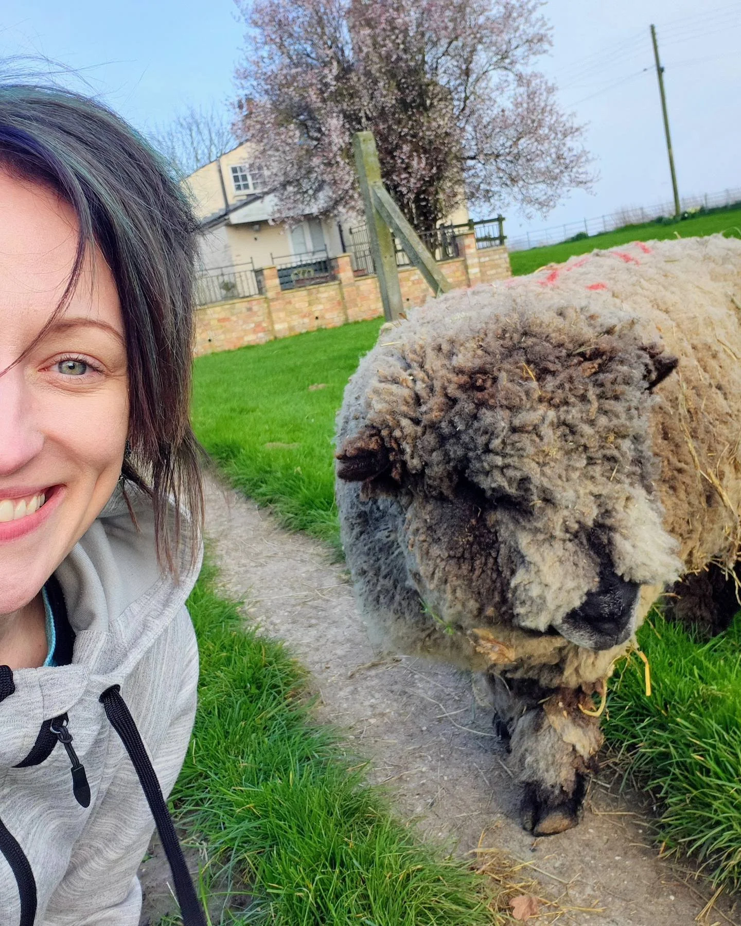 A woman taking a selfie with a dog on a grassy path in a rural area, with trees and a house in the background.