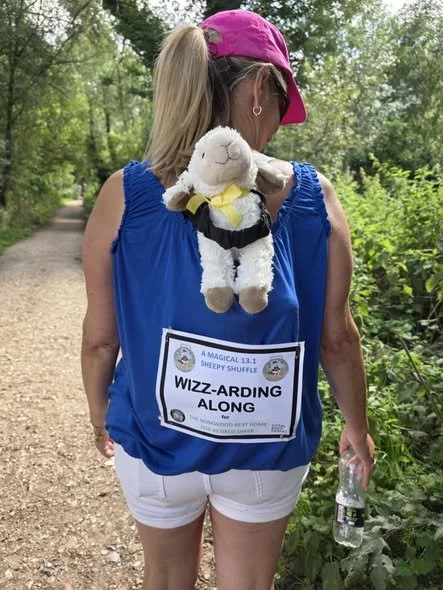 Woman with a puppy-shaped backpack on a trail in a wooded area, wearing a pink cap, blue sleeveless top, and white shorts, holding a water bottle.