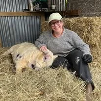 A woman sitting on haybales with a piglet, smiling at the camera.