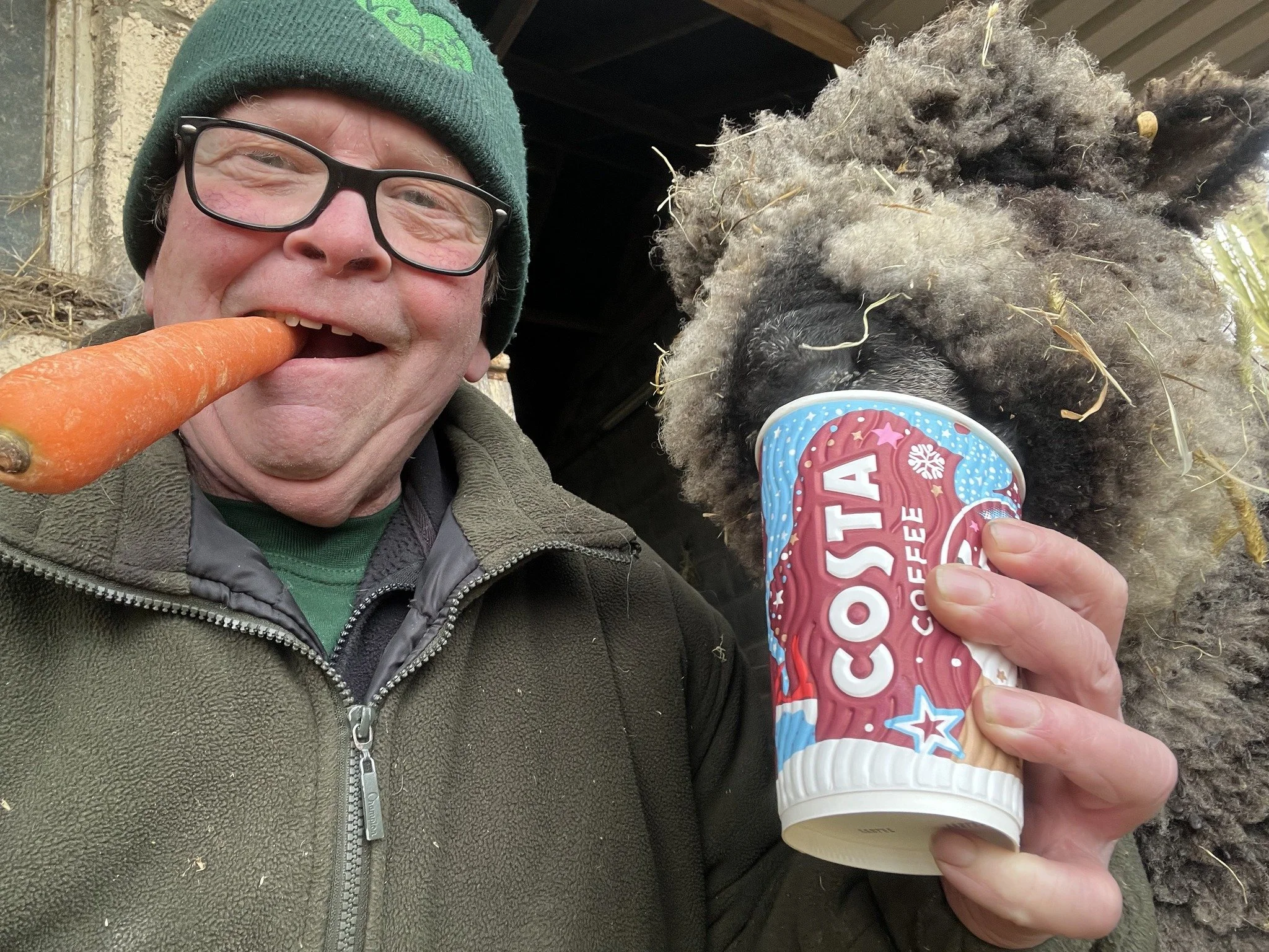 A man smiling, holding a carrot in his mouth and a cup of COSTA coffee, with a large black dog with curly fur in the background. The man is wearing glasses, a green beanie, and a green jacket.