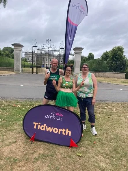 Three people standing outdoors in front of a gated estate, holding peace signs. A sign and flag promote the Tidworth park run event.