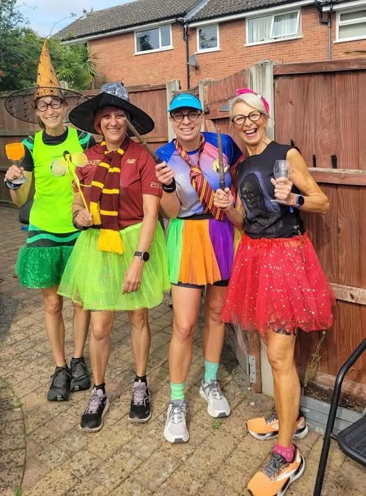 Four women dressed in colorful, whimsical costumes standing outdoors in a backyard, smiling and holding glasses. They are wearing tutus, hats, and accessories, indicating a fun, festive event.