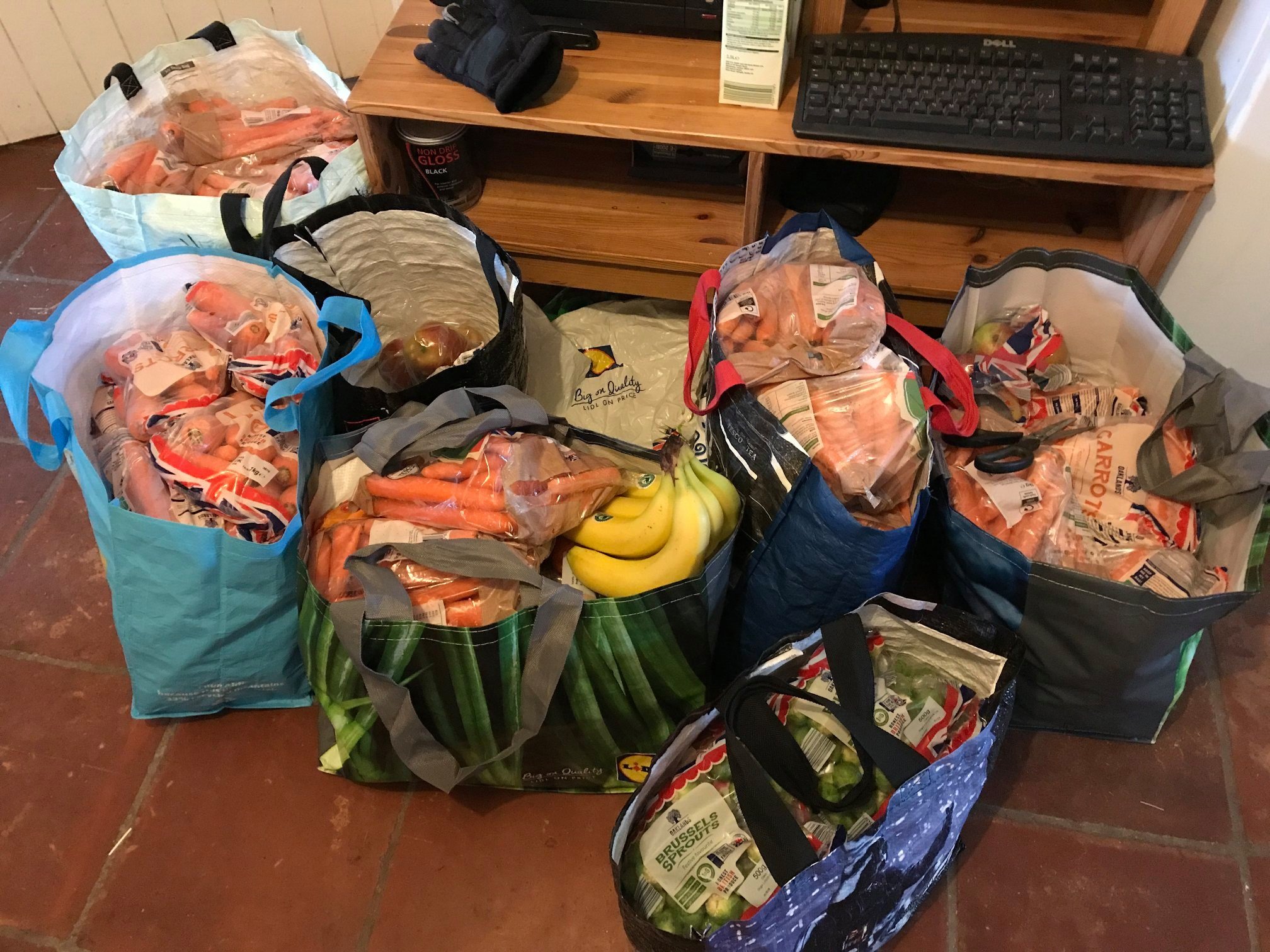 Multiple grocery bags filled with vegetables such as carrots, onions, bananas, and other produce, placed on a tiled floor in front of a wooden table with a keyboard on top.