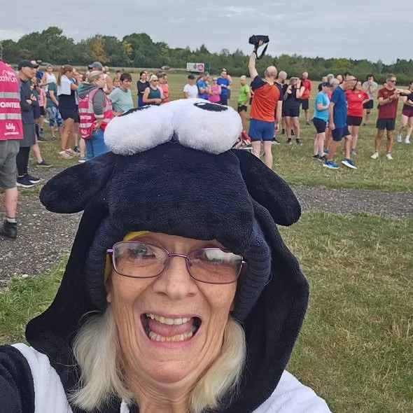 A woman taking a selfie at an outdoor event, wearing a hat with a sheep's head design, surrounded by a crowd of people on a grassy field.