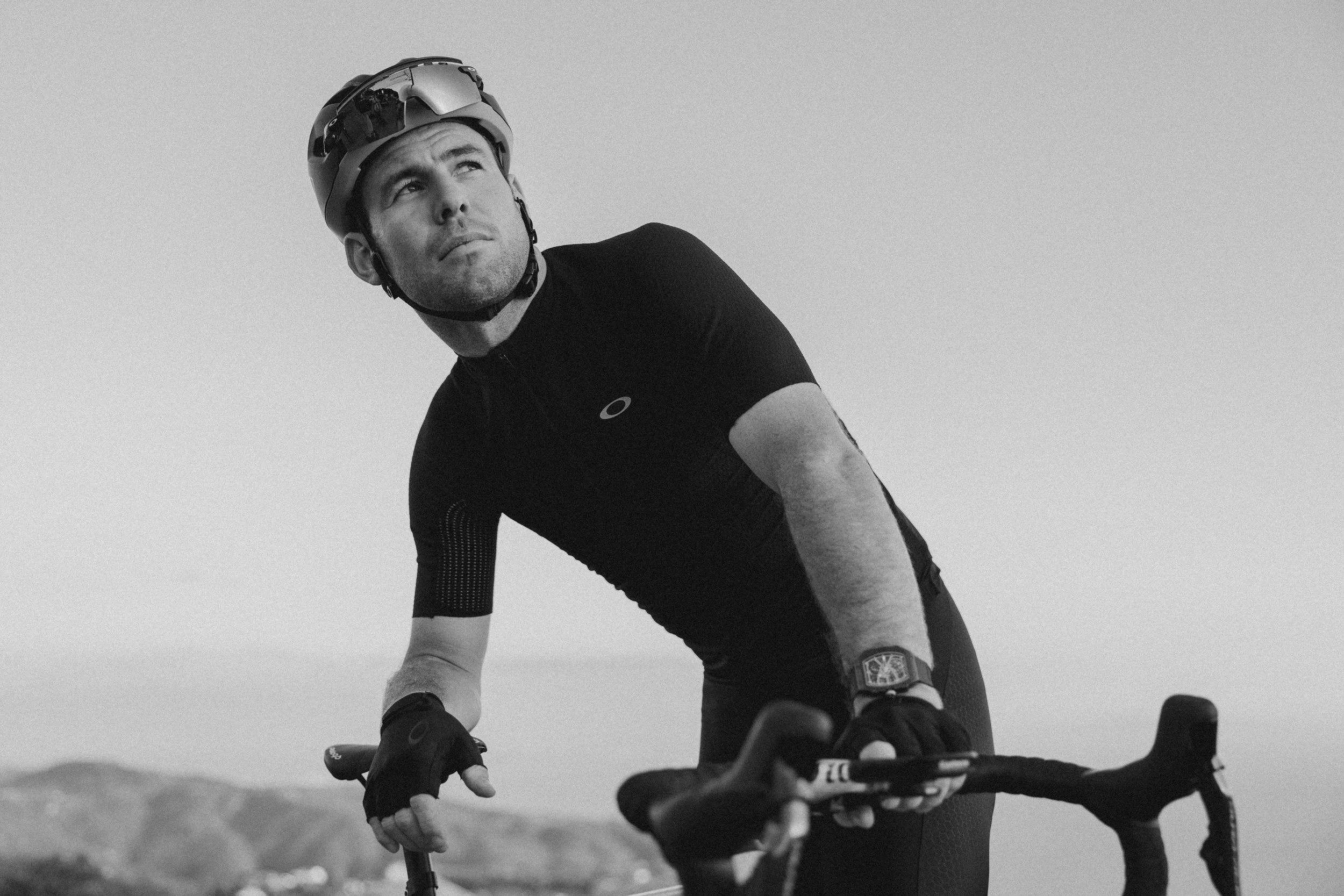 A man wearing a helmet and cycling gloves is leaning on his bicycle, looking upward with a serious expression. He is dressed in a black athletic shirt, and the background shows a landscape with hills under a clear sky.