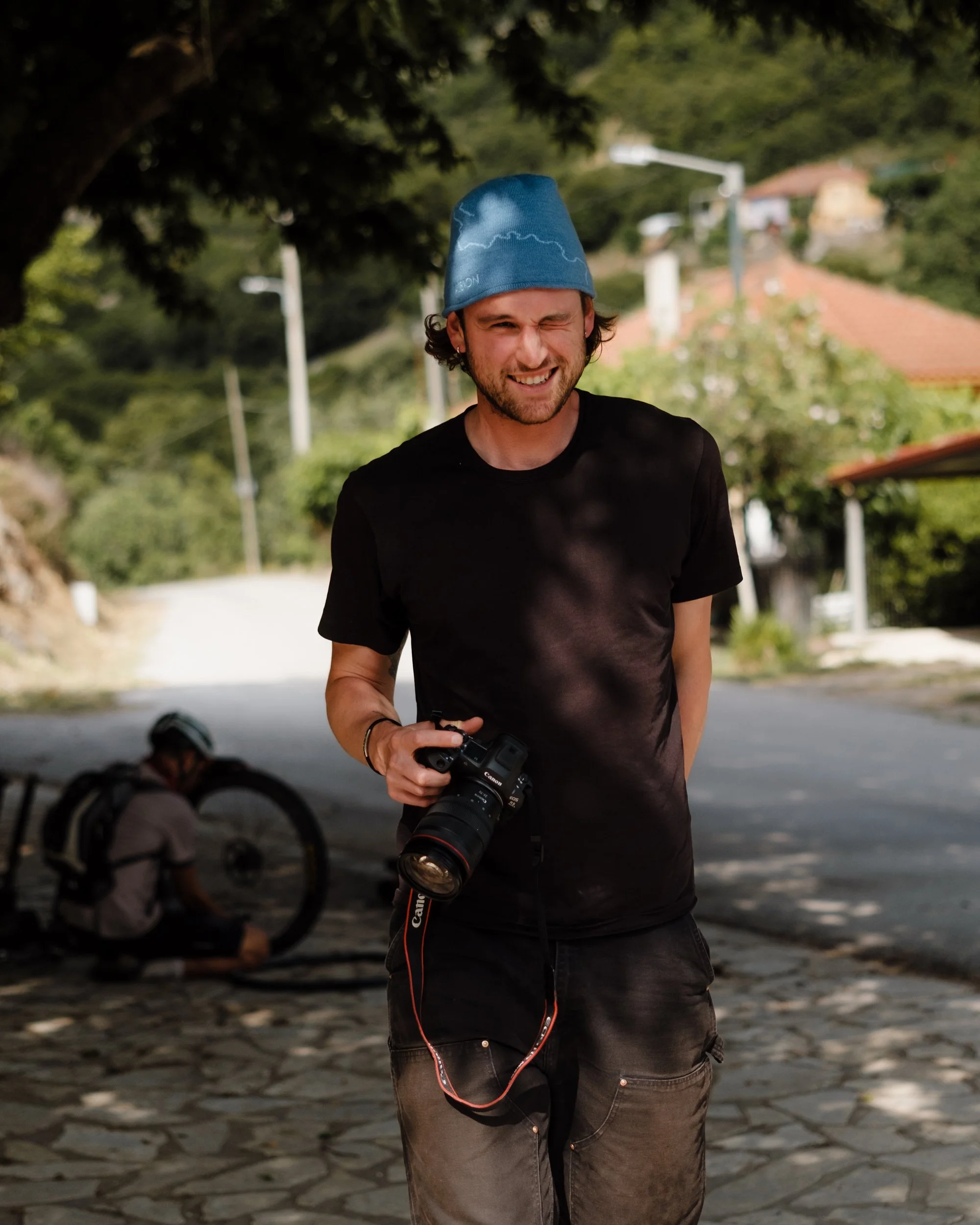 Young man with beard, wearing black t-shirt and blue beanie, smiling and holding a Canon camera outdoors, with a person on a bicycle in the background and trees and houses nearby.