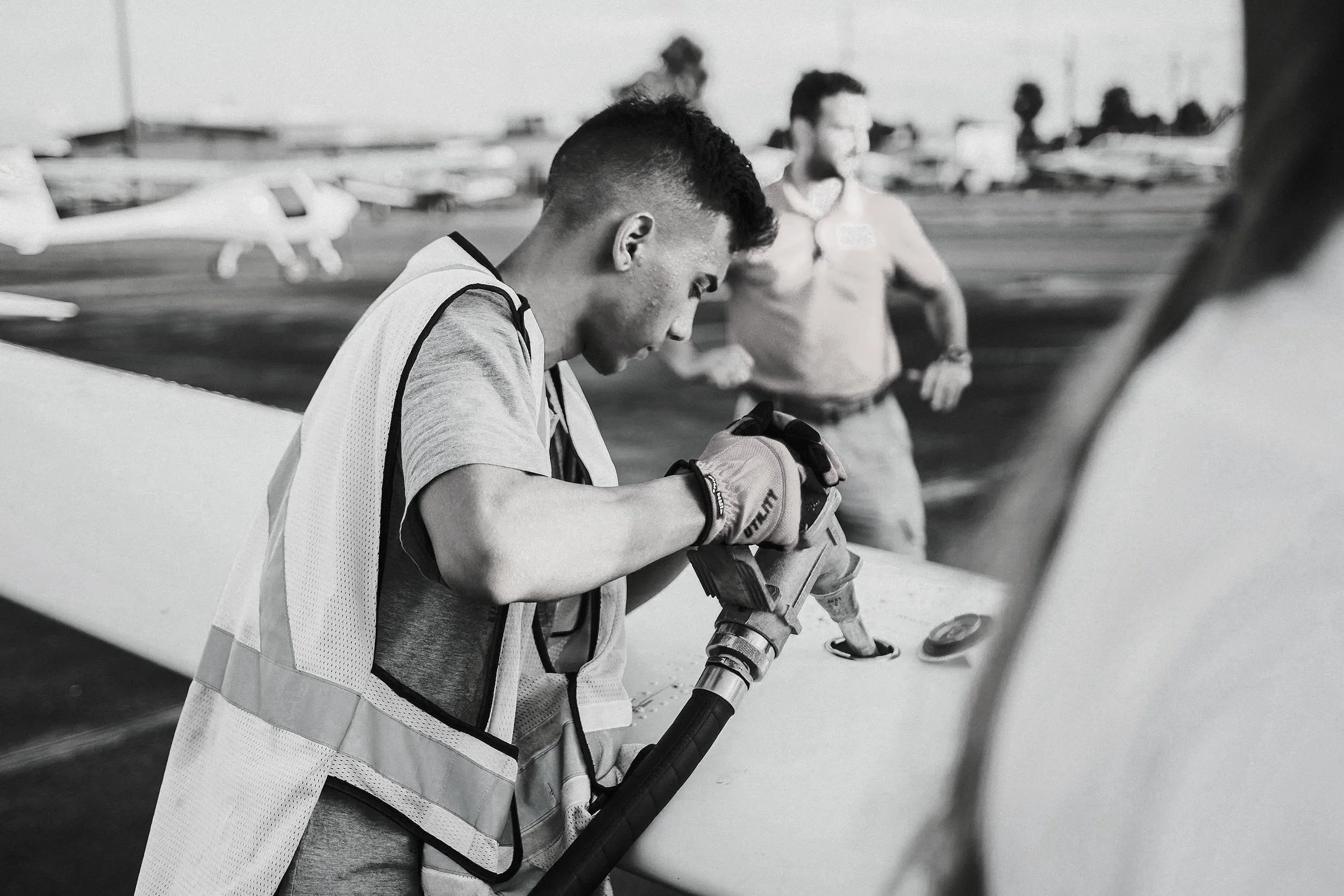 A young man in a sports vest, standing next to an aircraft's wing, is looking at his phone while wearing gloves. In the background, several small planes are parked on a tarmac at an airport, with blurred figures of other people and trees in the distance.